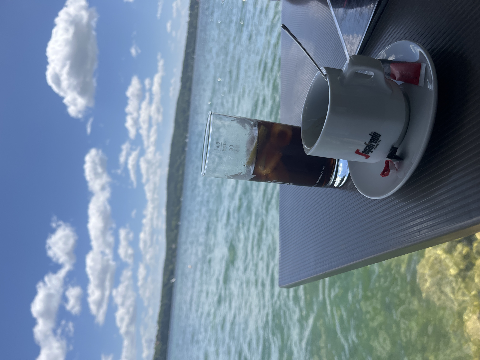 An outdoor cafe table is set with a white espresso cup and saucer, a glass of iced beverage, and a spoon. The cup has a red and black logo with the text "Segafredo ZANETTI". A red sugar packet is on the saucer. The table is positioned next to a large body of water, likely the Starnberger See, given the hilly, tree-lined shore visible in the distance under a bright blue sky with scattered white clouds. The water is clear and green, with visible rocks and sand beneath the surface. The overall scene suggests a leisurely moment by the lake.