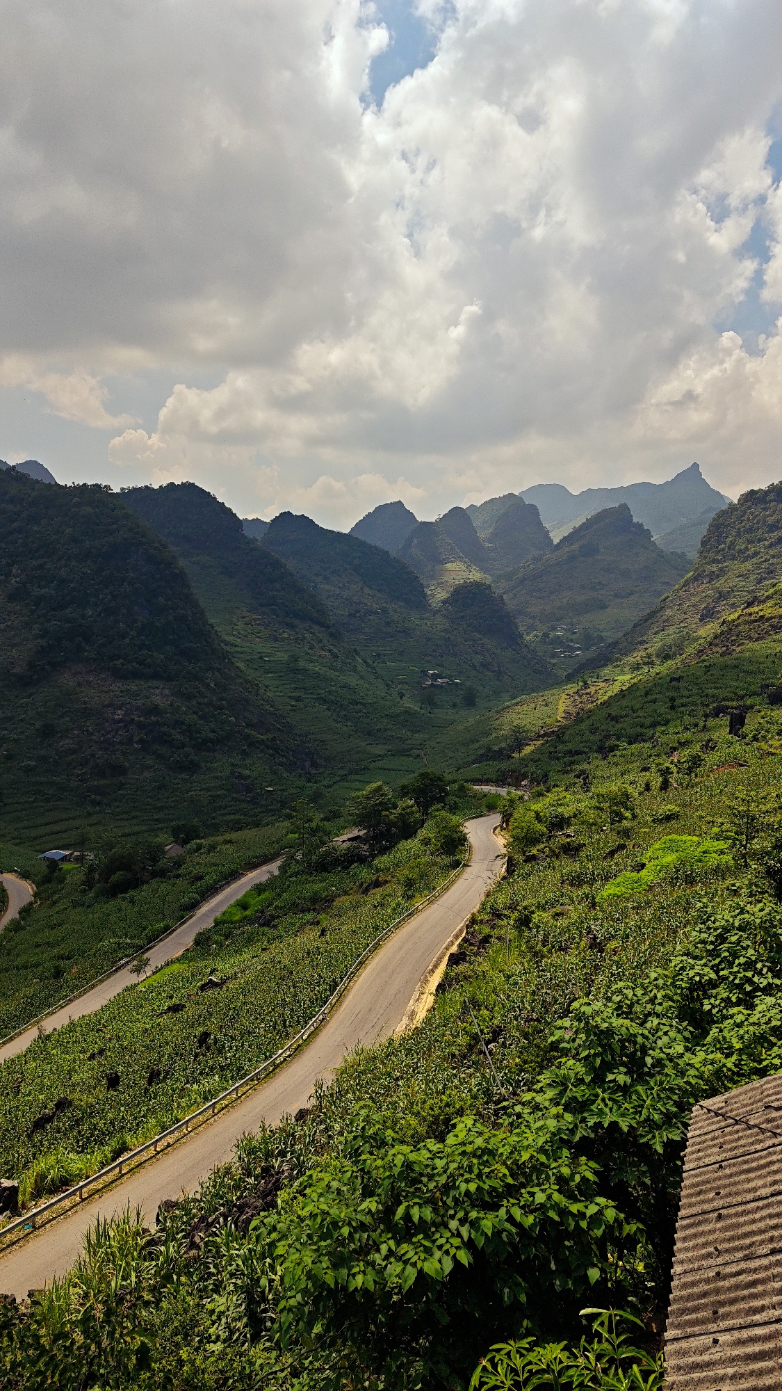 The image shows a winding road cutting through a mountainous region in Lũng Phìn Commune, Vietnam. The road is asphalt and has metal guardrails. The mountains are covered in lush, green vegetation. Some buildings are visible in the distance. The sky is cloudy. The view is from a high vantage point. A section of a corrugated roof is visible in the lower right corner.