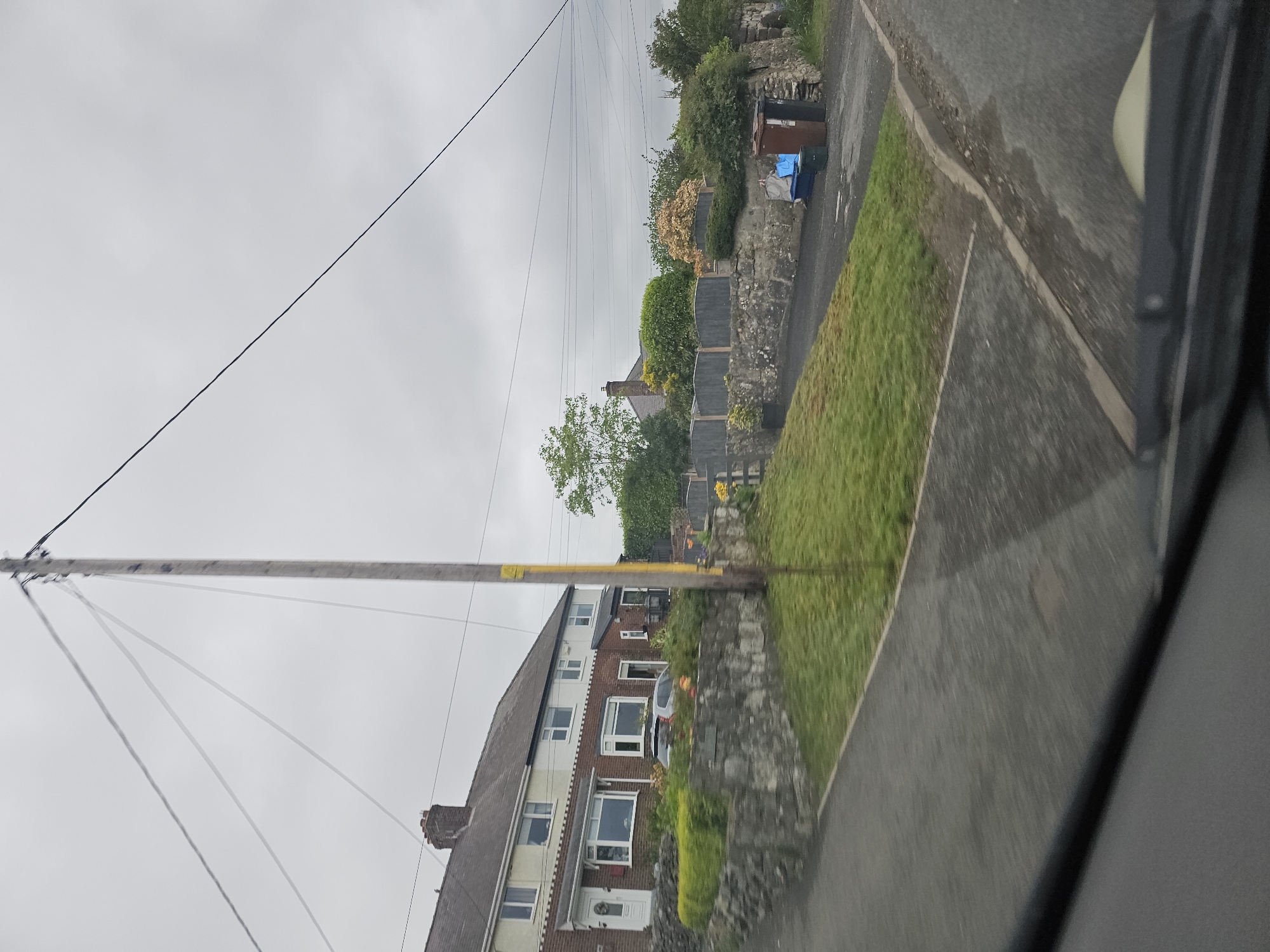 The image depicts a residential street scene in Halkyn, United Kingdom, taken from inside a vehicle. The perspective is slightly tilted. On the left side of the street, there are terraced houses constructed from brick and rendered walls. The roofs are pitched and predominantly dark brown. A wooden utility pole with multiple power lines is visible, crossing the street diagonally. On the right side of the street is a stone wall bordering properties, with various bushes and plants growing above it. Several wheelie bins are situated near the wall. The road surface is gray asphalt with some visible water marks. The sky is overcast and gray. The interior of the vehicle is visible on the right side of the image, with a portion of the window frame in view.