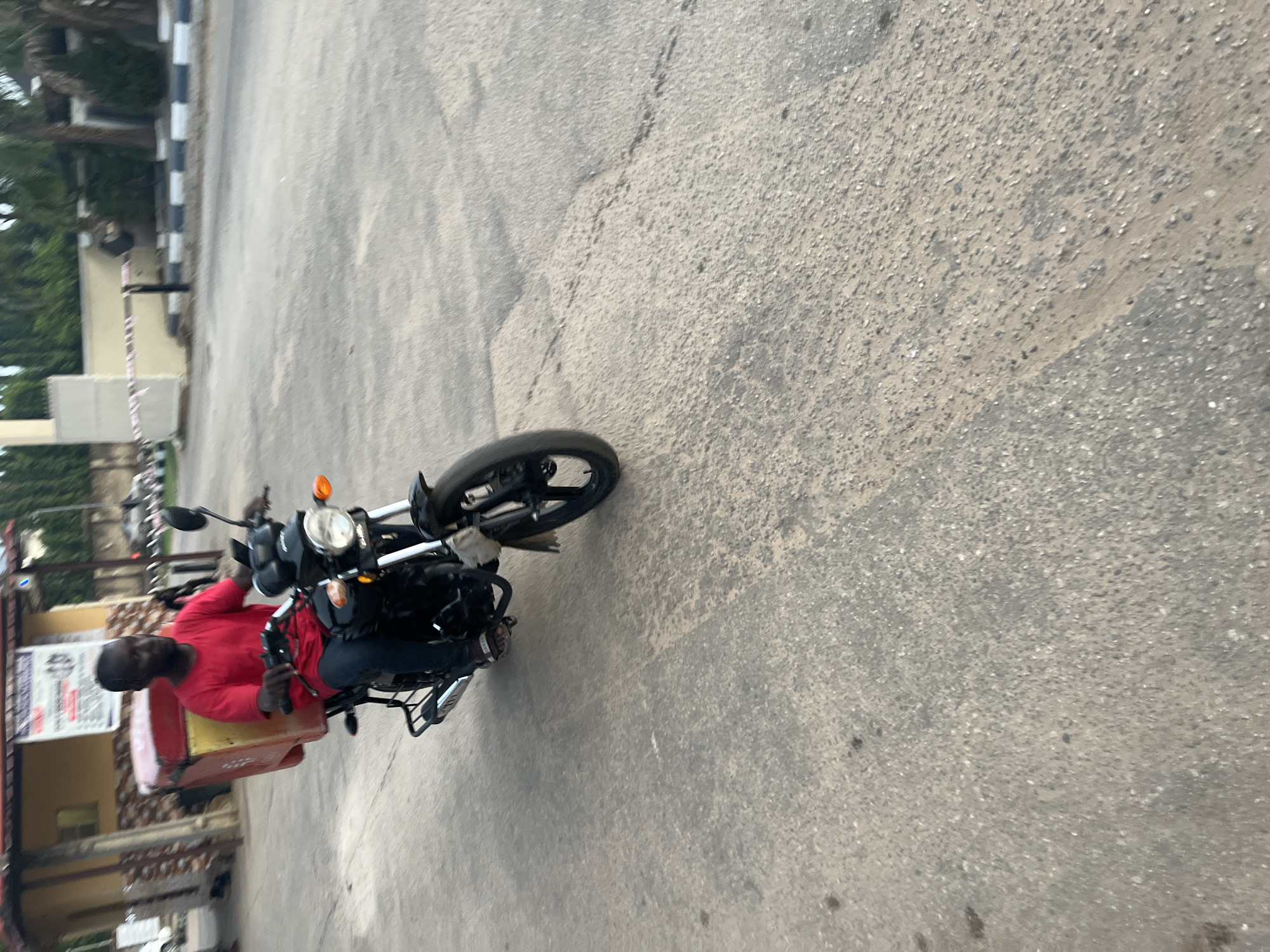 A man in a red shirt rides a black motorcycle with a red box attached to the back on an asphalt road. The location appears to be in a city, possibly Abuja, Nigeria, based on the architecture and road markings visible in the background. The road is cracked and uneven. The man is wearing blue jeans.