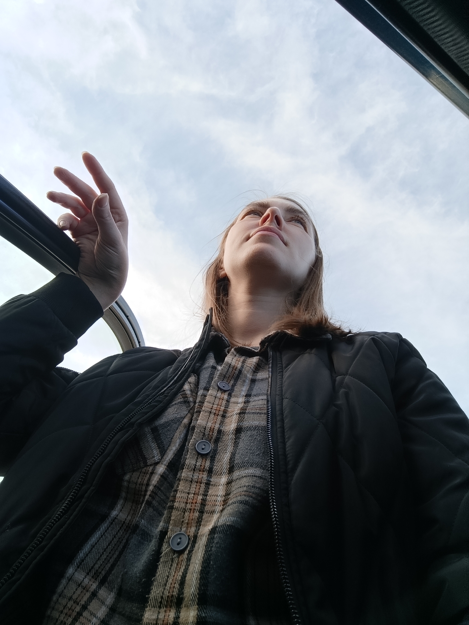 A low-angle shot captures a person with light skin and shoulder-length brown hair, looking upwards towards the sky. The person is wearing a buttoned, plaid shirt under a black quilted jacket. Their left hand is partially visible, holding onto a dark metallic frame. The backdrop is a cloudy sky, suggesting daylight. A section of a dark-colored vehicle interior is visible at the top right of the frame.
There is nothing in this image to indicate the location as the city of Chaikovsky, Russia.