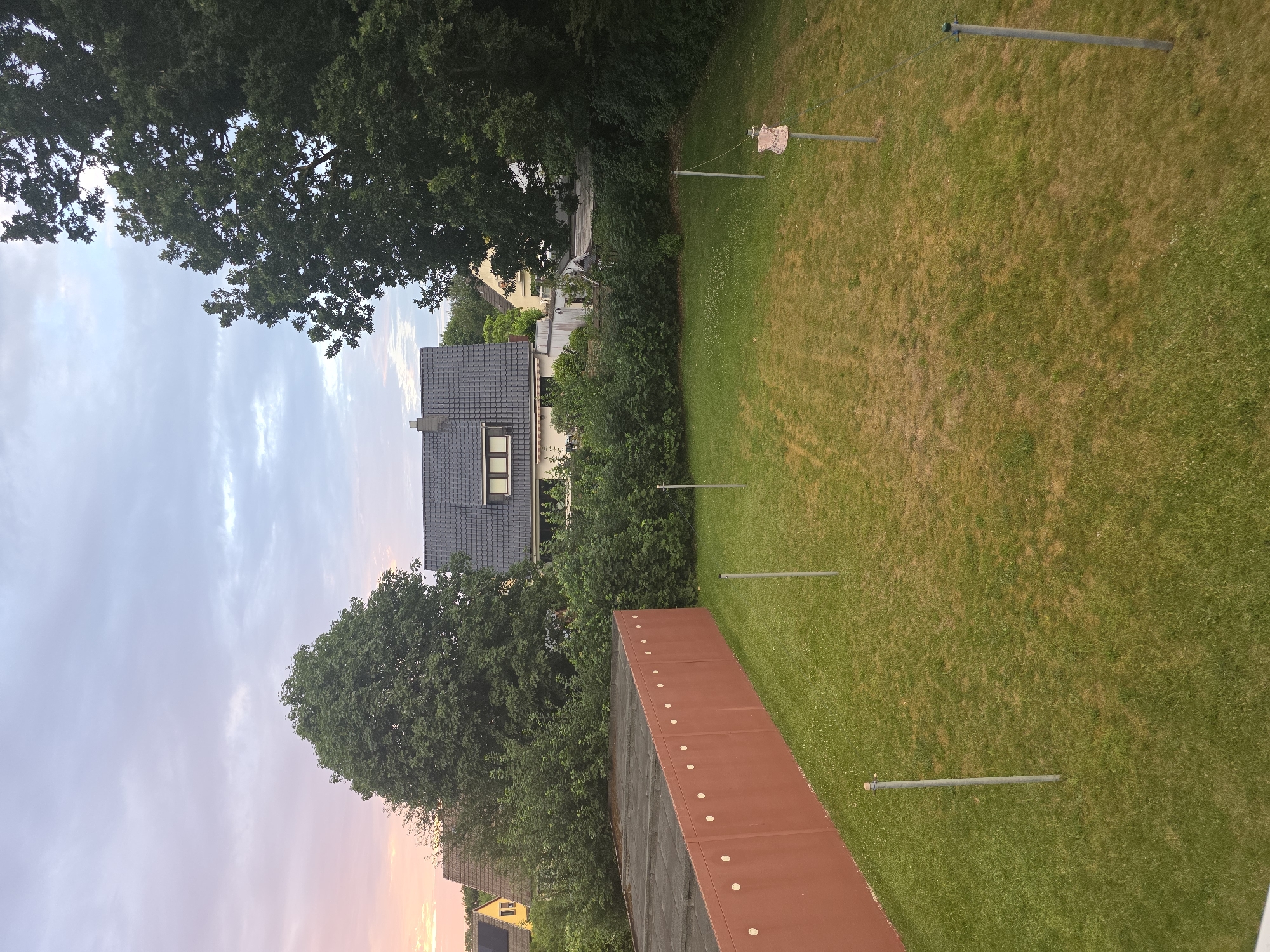 The image depicts a residential backyard in Uetersen, Germany, at dusk. A red, concrete wall with circular indentations runs along the left side of the frame. The backyard itself is covered in grass, with several metal poles spaced across it, some of which appear to be clothesline supports. One pole in the middle right has a decorative, pink object resembling a dress or garment hanging from it. In the background, there are several houses with pitched roofs, surrounded by lush green trees and bushes. The sky is cloudy, with soft hues of pink and orange on the left side, suggesting the setting sun. The overall atmosphere is peaceful and suburban.
