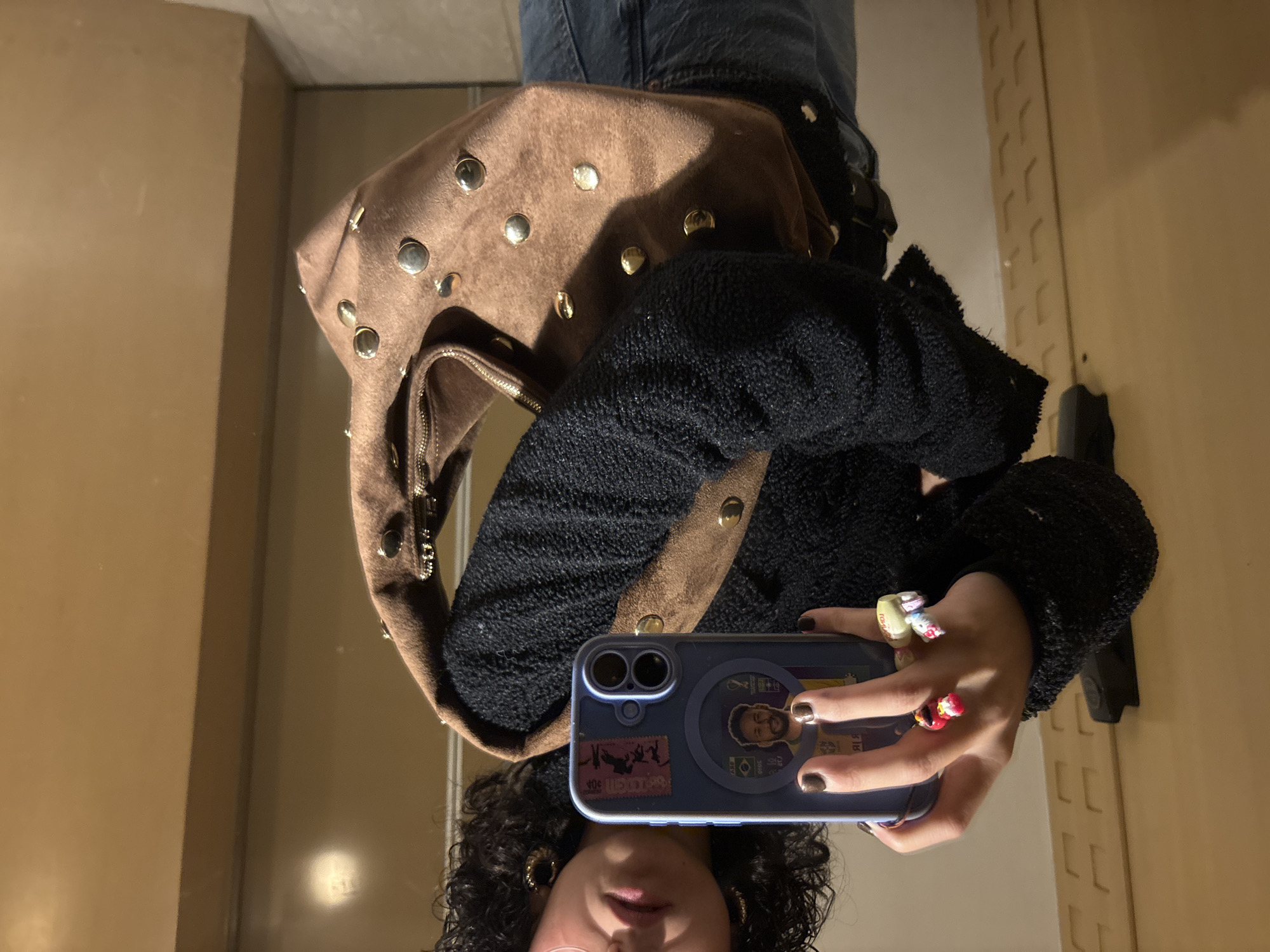 A person is taking a selfie in a mirror. The person is wearing jeans, a black sweater with brown trim, and carrying a brown bag with metal studs. The person is holding a blue smartphone with stickers on the back, and wearing two colorful rings on their fingers. The mirror reflects a beige wall with vertical slots. The photograph was taken inside, in a location that may be in Mexico City, Mexico.