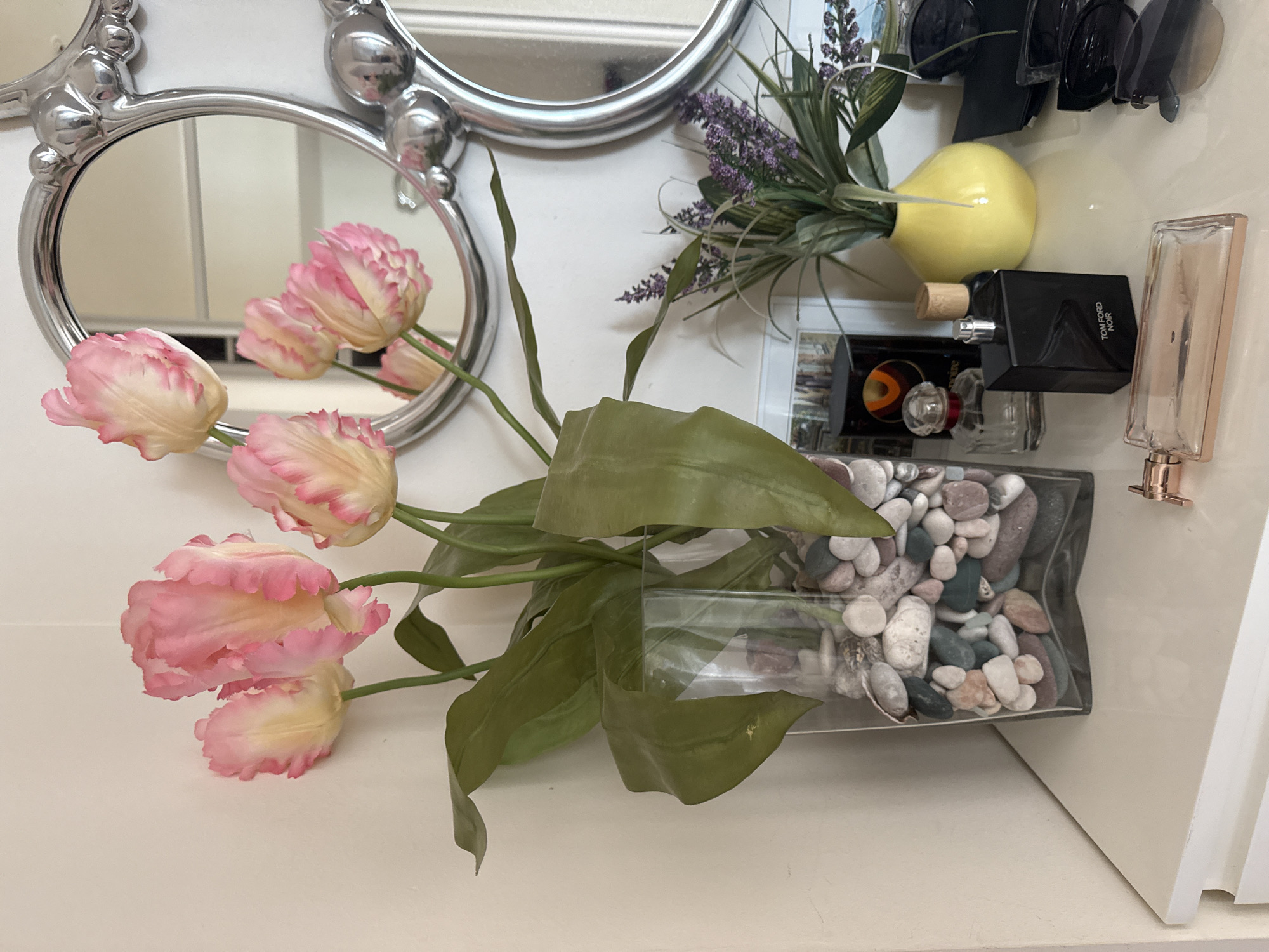A still life composition features a vase of pink and yellow ruffled tulips on a white surface, possibly a dresser or shelf. The vase is clear glass filled with decorative pebbles. To the right of the tulips, a decorative mirror with a silver ornate frame is mounted on a wall, reflecting the tulips and part of the room. In front of the mirror, several objects are arranged. These include two perfume bottles, one black and rectangular with "TOM FORD NOIR" printed on it, and a smaller clear bottle with a red cap. Behind them, a small yellow vase holds a bouquet of lavender and green foliage. A pair of sunglasses rests to the far right. A rose gold colored perfume bottle lies horizontally in the foreground. The scene is well-lit, with soft shadows. No people are visible. The specific location in Budva, Montenegro, cannot be determined from the visual information provided in the image.