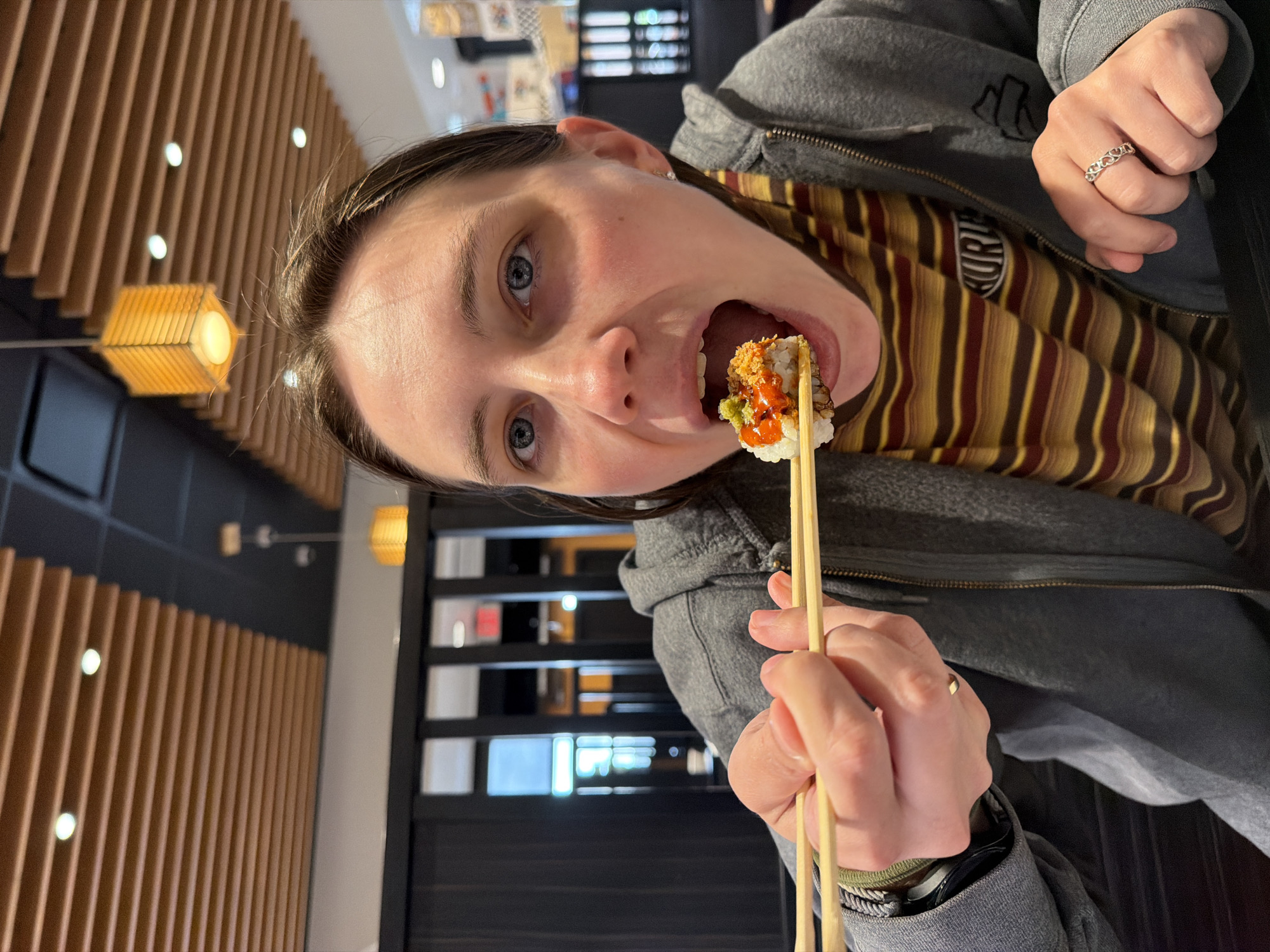 A young woman with blue eyes and light brown hair is shown from the chest up, in a medium close-up shot, as she is about to eat a piece of sushi with chopsticks. She is wearing a gray zip-up hoodie over a horizontally striped shirt in brown, yellow, and maroon. Her mouth is open as she brings the sushi towards it. The sushi is held by a pair of wooden chopsticks, and it appears to be topped with a spicy sauce and some green and brown toppings. A ring with a heart pattern is visible on her right hand. The background shows a restaurant interior with wooden slatted ceilings and hanging lamps. The scene appears to be in a restaurant in Beaverton, Oregon, USA.