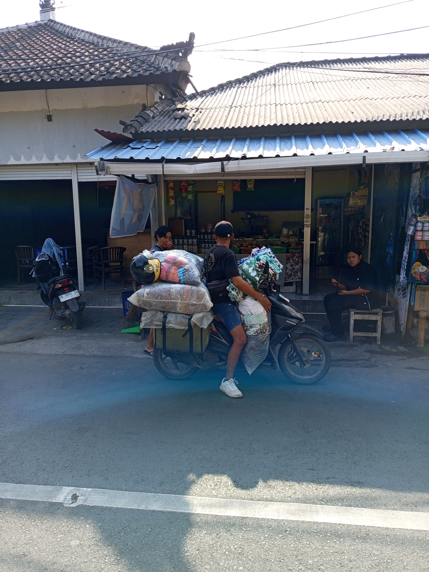 The image shows a street scene in Denpasar, Indonesia, with a focus on a man riding a motorcycle overloaded with goods. The man, wearing a black t-shirt, denim shorts, and a baseball cap, is seated on a black motorcycle. His legs straddle the motorcycle, and he is holding onto handlebars. The motorcycle is heavily laden with various items, including large plastic-wrapped bundles and what appear to be textiles or clothing. A yellow and black helmet is placed on top of one of the bundles.
Behind the motorcycle, a building with a corrugated metal roof and a traditional tiled roof extension is visible. The building appears to be a shop or market stall with an open front, displaying shelves of goods and various items for sale. A woman in a black shirt is seated on a small wooden stool outside the shop, holding a mobile phone. Another motorcycle is parked to the left of the scene, covered with a blue cloth. The street is paved with asphalt and has a white painted line running across it. The overall impression is that of a busy commercial area in Denpasar, with daily commerce taking place.