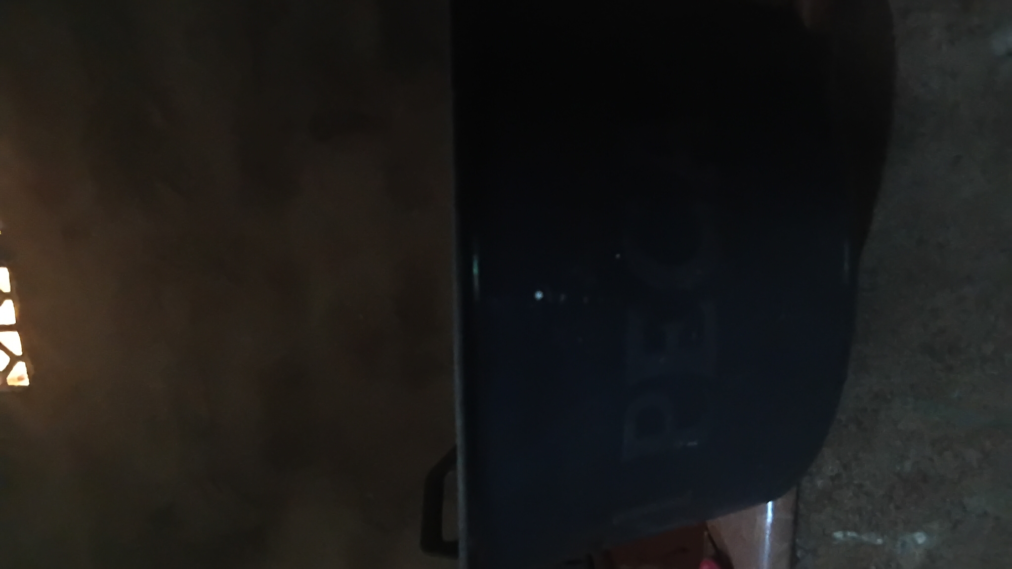 A black plastic tub with handles is shown in a dark, indoor setting. The tub has faint white lettering on its side, which appears to read "APEC". The background consists of a dark, textured wall with a patterned window or vent at the top. The floor is a rough, concrete-like surface. The lighting is dim, casting shadows and obscuring finer details. This scene is likely in a residential or utility area in Prabumulih, Indonesia.