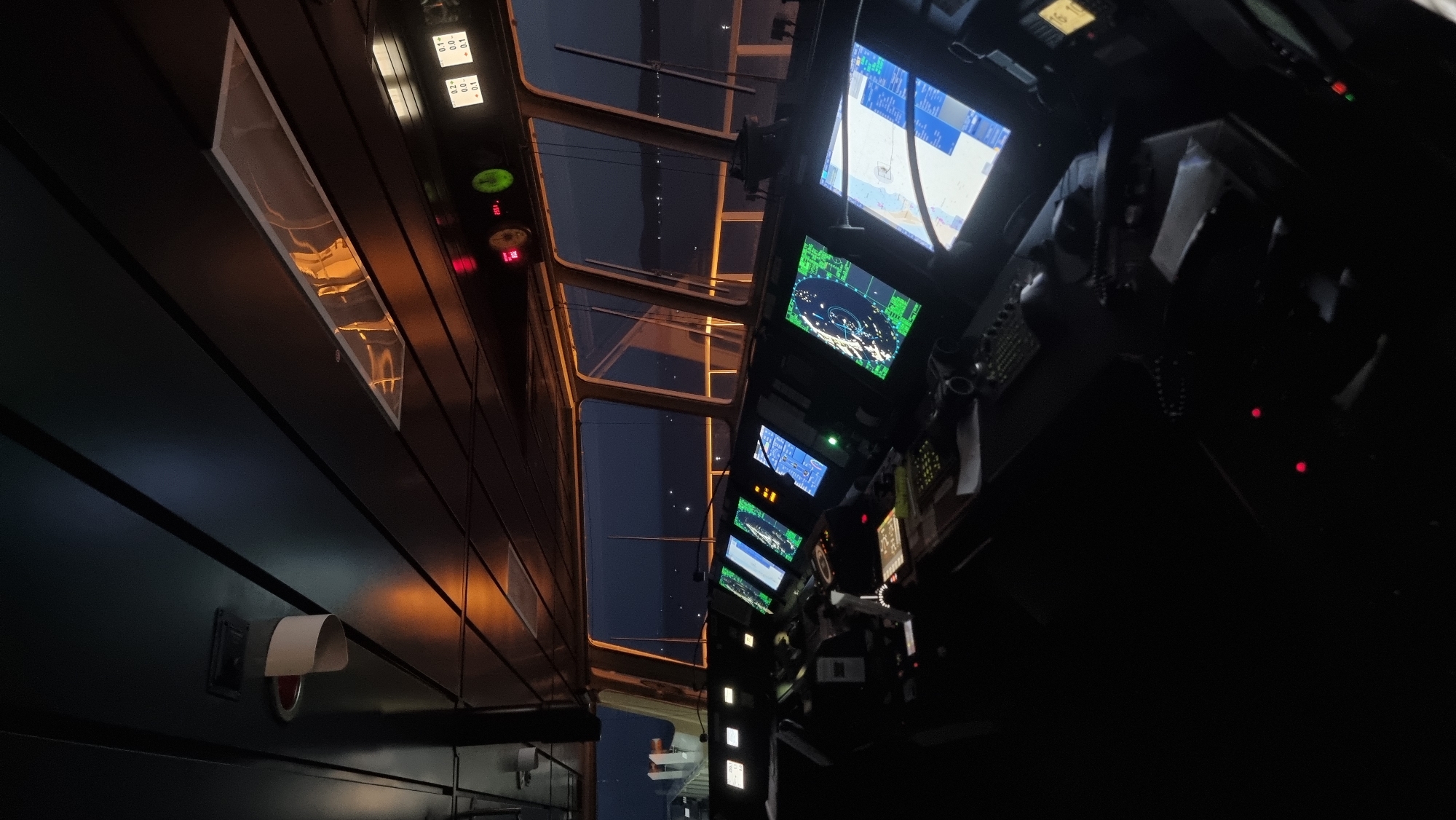 The image depicts the interior of a ship's bridge at dusk or night. The main focus is on a console filled with multiple electronic displays, most of which are showing navigational charts and radar readouts in green and blue. The displays are arranged in a semi-circular fashion, suggesting the command center of a vessel. Outside the large windows, the dark sea and sky are visible, with distant lights possibly indicating land or other ships. The bridge is dimly lit by the glow of the screens and some overhead lighting, creating a professional and functional atmosphere. The scene is devoid of people, and the equipment appears to be in operational condition. No specific details are visible to place this scene in Trincomalee, Sri Lanka, as the bridge of a ship is a standardized environment.