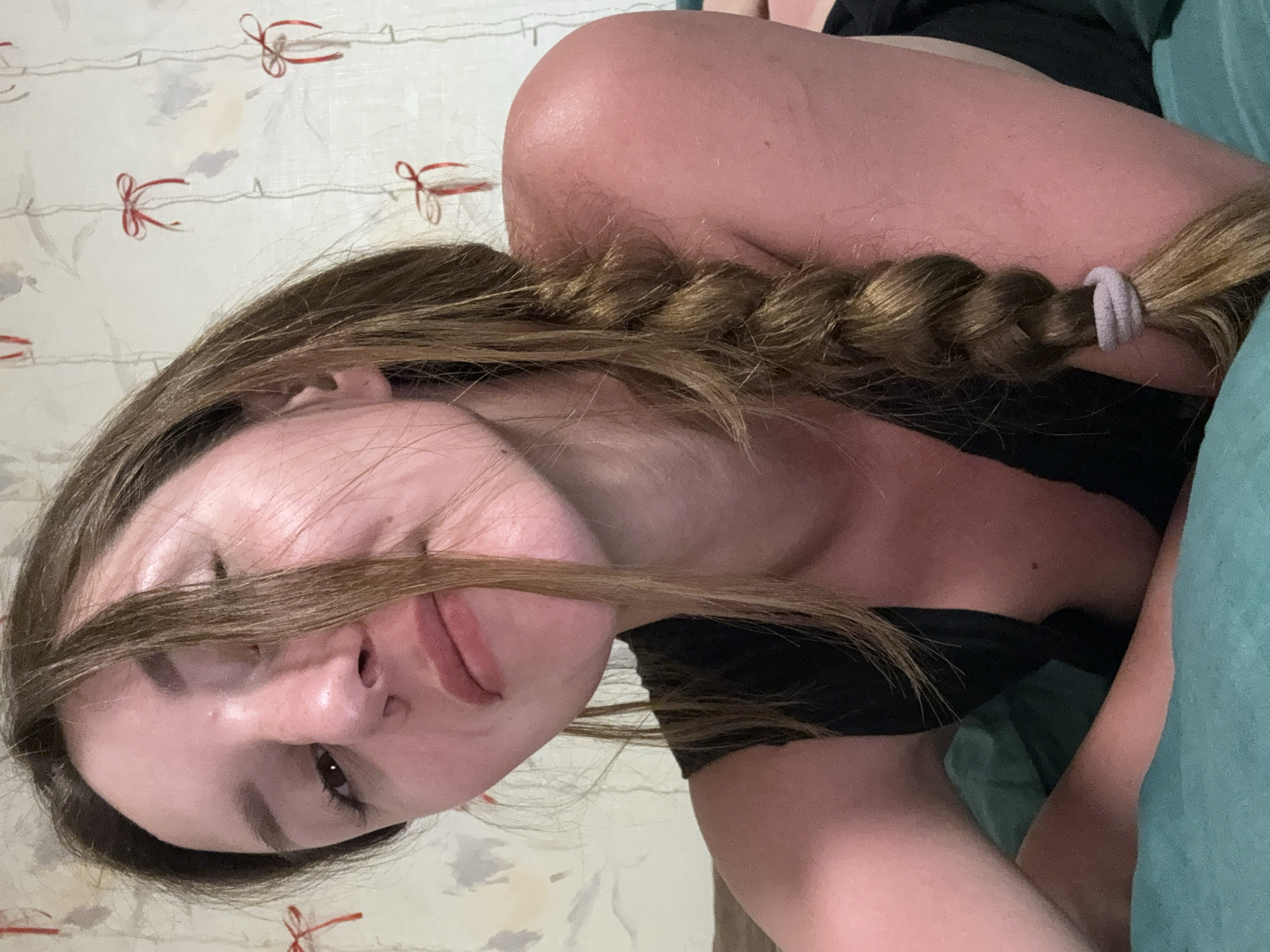 The image shows a close-up, low-angle shot of a young woman lying on her side on a teal-colored surface, possibly a bed or sofa. She has long, brown hair, mostly falling over her face, with a braid visible down her back. She is wearing a black top. Her skin appears reddish, suggesting a sunburn. The background is a wall with a patterned wallpaper featuring delicate red bows and vertical lines. The scene is indoors, and the lighting is soft. No specific location in Zhodzina, Belarus is identifiable from the image.