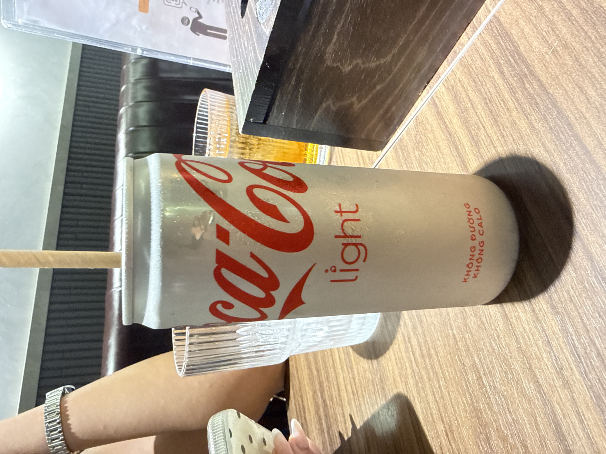 The image depicts a Coca-Cola Light can with a straw, resting on a wooden table next to a glass. Part of an individual's arm, adorned with a silver watch, is visible along with a portion of a shoe. The text on the can is in Vietnamese. The location is inferred to be Đà Nẵng, Vietnam, based on the beverage can's regional availability and language on the can.