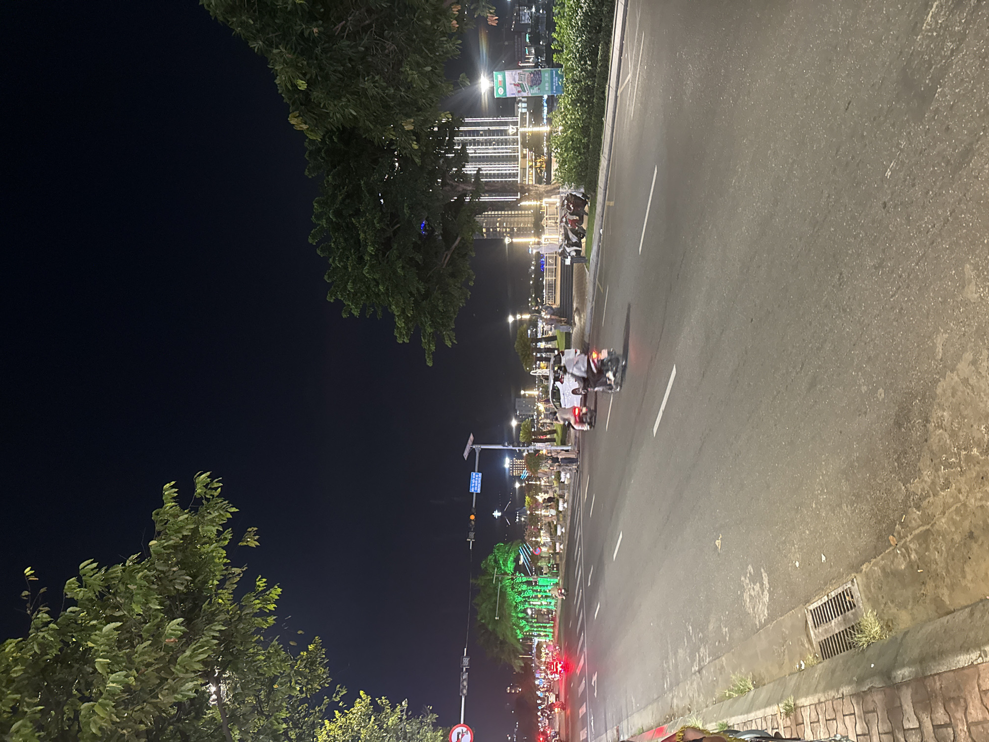 At night in an unknown city, an upward-angled view shows a well-lit multi-lane roadway with parked scooters and moving motorbikes. The road is lined with trees and green shrubbery. The backdrop features brightly lit skyscrapers, light posts, and decorative lights. A traffic light and traffic sign are visible at the bottom of the frame. A storm drain is visible in the bottom right corner. A few people are standing along the sidewalk.