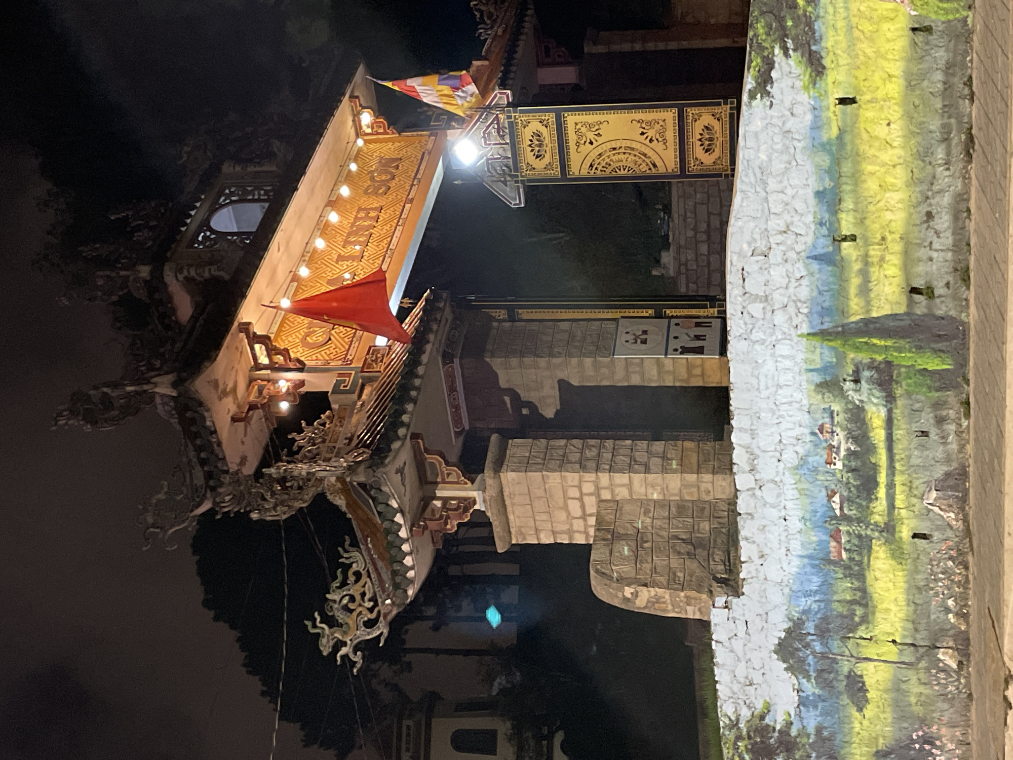 This is a nighttime photograph of the entrance to the Linh Son Pagoda in Da Lat, Vietnam. The entrance features a traditional Vietnamese architectural style with ornate roofing, decorative carvings, and intricate tilework. Two flags are visible; a Vietnamese flag on the left and a colorful Buddhist flag on the right. The pagoda is illuminated by several strings of lights. In the foreground, a mural adorns a low wall, depicting a pastoral landscape with houses, trees, and a field of yellow flowers. The overall scene conveys a sense of cultural and religious significance, set against a dark, quiet night sky.