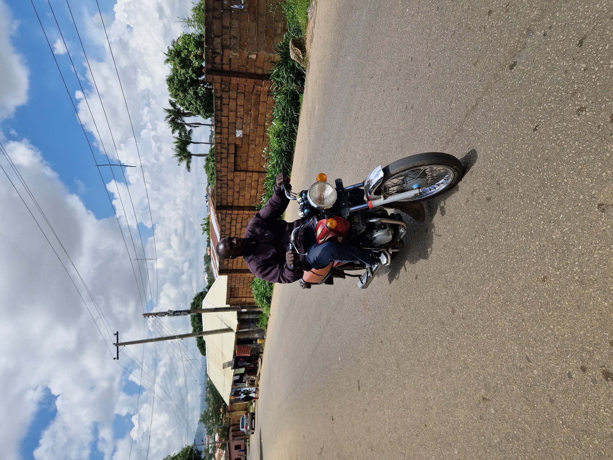 A full shot, eye-level, outdoor, natural light, medium shot captures a man riding a motorcycle on a paved road. The man, wearing a dark jacket, blue jeans, and white sneakers, is seated on a red motorcycle with a tan seat and chrome accents. He is looking to his right as he rides forward. The road is lined with a brick wall and lush green vegetation on the right side. In the background, there are palm trees, buildings with light-colored roofs, a power pole with numerous wires, and a blue sky with white clouds. A silver car is visible in the distance on the left side of the road, and a few pedestrians are near the buildings. The overall scene depicts a common urban or suburban street environment.