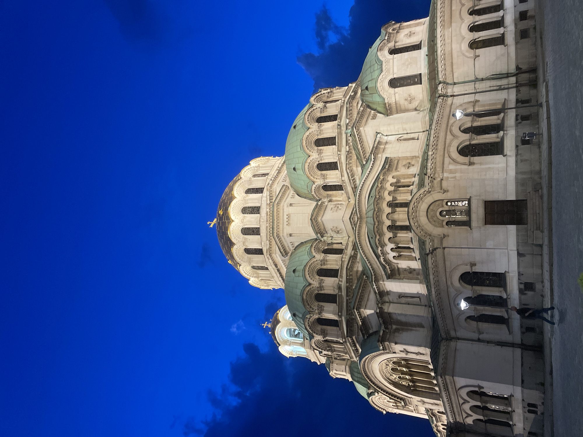 The image shows the Alexander Nevsky Cathedral in Sofia, Bulgaria, at night. The cathedral's domes are illuminated, contrasting with the dark blue sky. A person is walking along the sidewalk near the base of the building.
