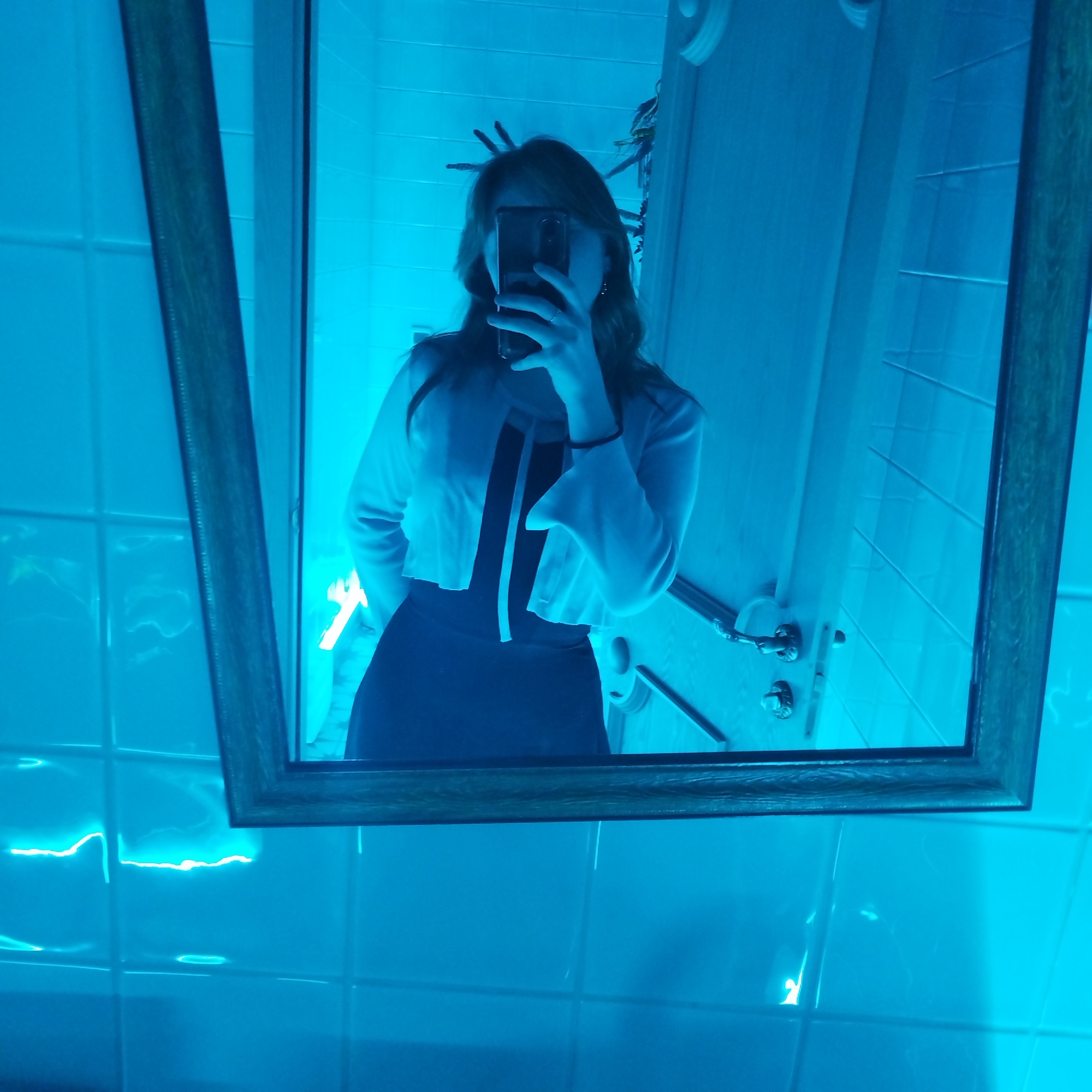 A mirror selfie, taken in Yekaterinburg, Russia, features a person within an indoor setting, likely a bathroom, intensely illuminated by blue light. The subject, a female with long hair, is visible from the waist up, holding a smartphone to obscure her face while capturing her reflection. She is dressed in a black outfit, possibly a dress, topped with a light-colored, cropped cardigan featuring wide, bell-shaped sleeves. A ring is visible on her right hand.
The photo is framed by a rectangular mirror with a wooden border. The reflection in the mirror reveals a tiled wall behind the subject, characteristic of a bathroom. Partially visible on the right side of the reflection are elements of a door, including a handle. A bright, neon-like line of blue light, likely from an LED strip, reflects distinctly on the tiled surface in the bottom left area of the image. The pervasive blue lighting creates a strong monochromatic effect, saturating the entire scene in cool tones and making other colors appear subdued. There is no visible text in the image.