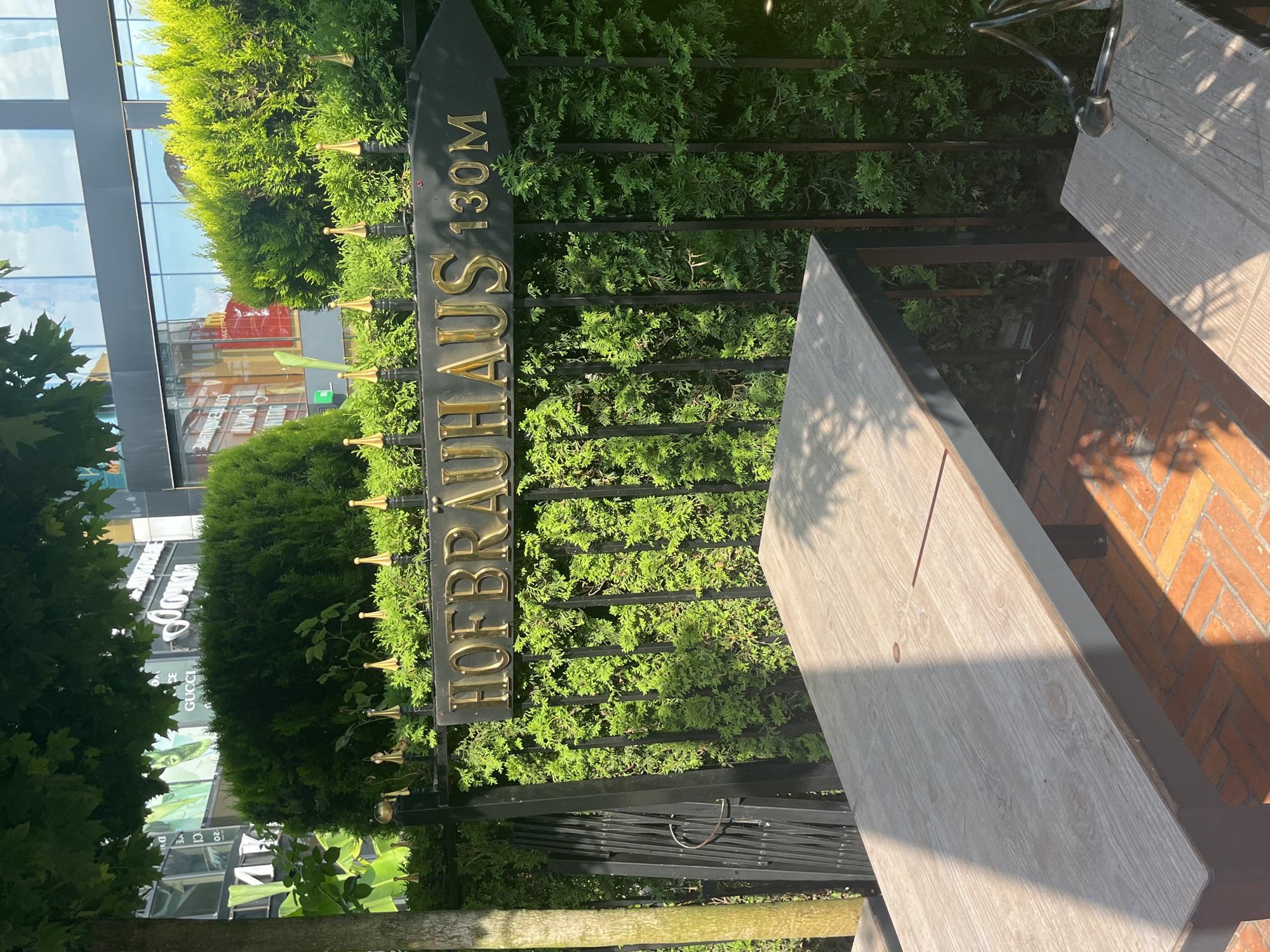 This image shows an outdoor seating area with a large wooden table in the foreground. The table is positioned on a brick patio, with a black metal fence and lush green hedges behind it. A black sign with gold lettering points to the right, reading "HOFBRAUHAUS 130M". The background shows a modern building with large glass windows, reflecting the sky and surrounding trees. The lighting suggests it is daytime, with sunlight casting shadows on the table and patio. The scene appears to be in an urban environment, possibly a restaurant or beer garden. The overall impression is one of a pleasant outdoor dining or lounging space.