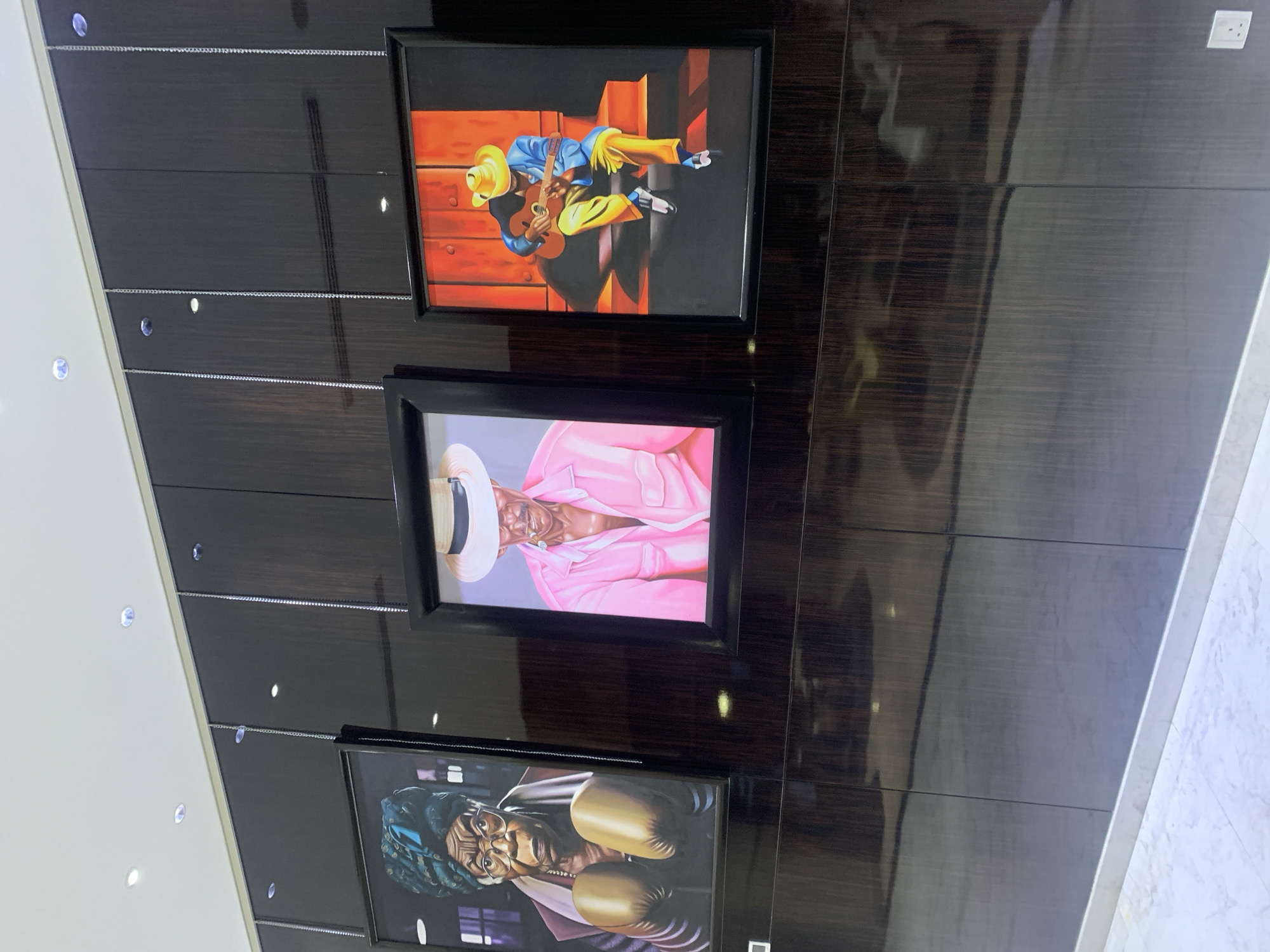 The image shows a wall in what appears to be a hallway or interior space in Dar es-Salaam, Tanzania. The wall is covered in dark wood paneling with horizontal seams and reflective surfaces. Three framed pictures hang vertically along the wall. The topmost picture depicts a person playing a guitar against a warm-toned background. The middle picture portrays a person in a pink outfit and hat. The bottom picture shows a person wearing boxing gloves. Small, round lighting fixtures are visible on the left side of the wall. On the right side of the wall is white marble. A white electrical outlet is visible near the top right corner of the frame. The scene appears to be indoors in a building of modern design.
