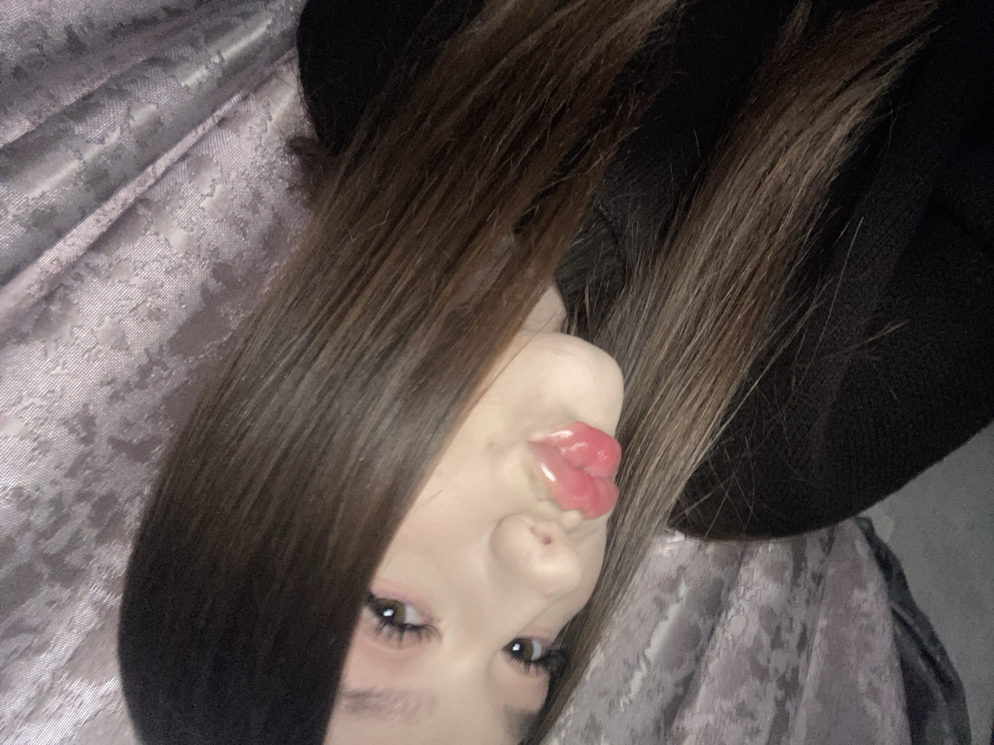 The image is a close-up portrait of a person, likely a young woman, presented rotated ninety degrees clockwise. The main subject's face is visible in the lower right portion of the frame, with her long, straight dark brown hair flowing across the image, framing her features.
She has dark brown eyes with prominent, long eyelashes and a hint of pink eyeshadow on her eyelids. Her lips are puckered in a glossy, bright pink "kiss" or "duck face" expression, suggesting a playful or stylized pose. Her skin appears fair.
The background consists of a textured fabric, possibly a curtain or bedding, in a light lavender or silver-grey hue with a subtle woven pattern. This fabric occupies the left side of the image. A dark, solid area, which could be part of a garment, a hat, or a shadowed space, is visible on the right side of the frame, behind her hair.
The overall impression is that of a posed, indoor selfie. No specific time of day, weather conditions, or explicit location cues beyond an indoor setting are discernible from the image content. There is no visible text.