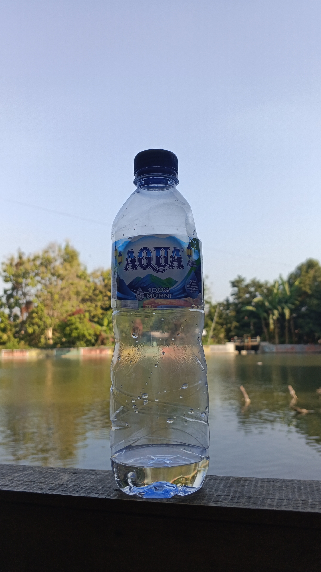 A plastic bottle of Aqua brand bottled water is placed on a wooden railing. The bottle is partially filled with water, and condensation is visible on its exterior. In the background, a body of water, possibly a lake or pond, is visible, with trees and a clear sky. The setting appears to be an outdoor location in Rengel, Indonesia, suggesting a recreational or natural area.