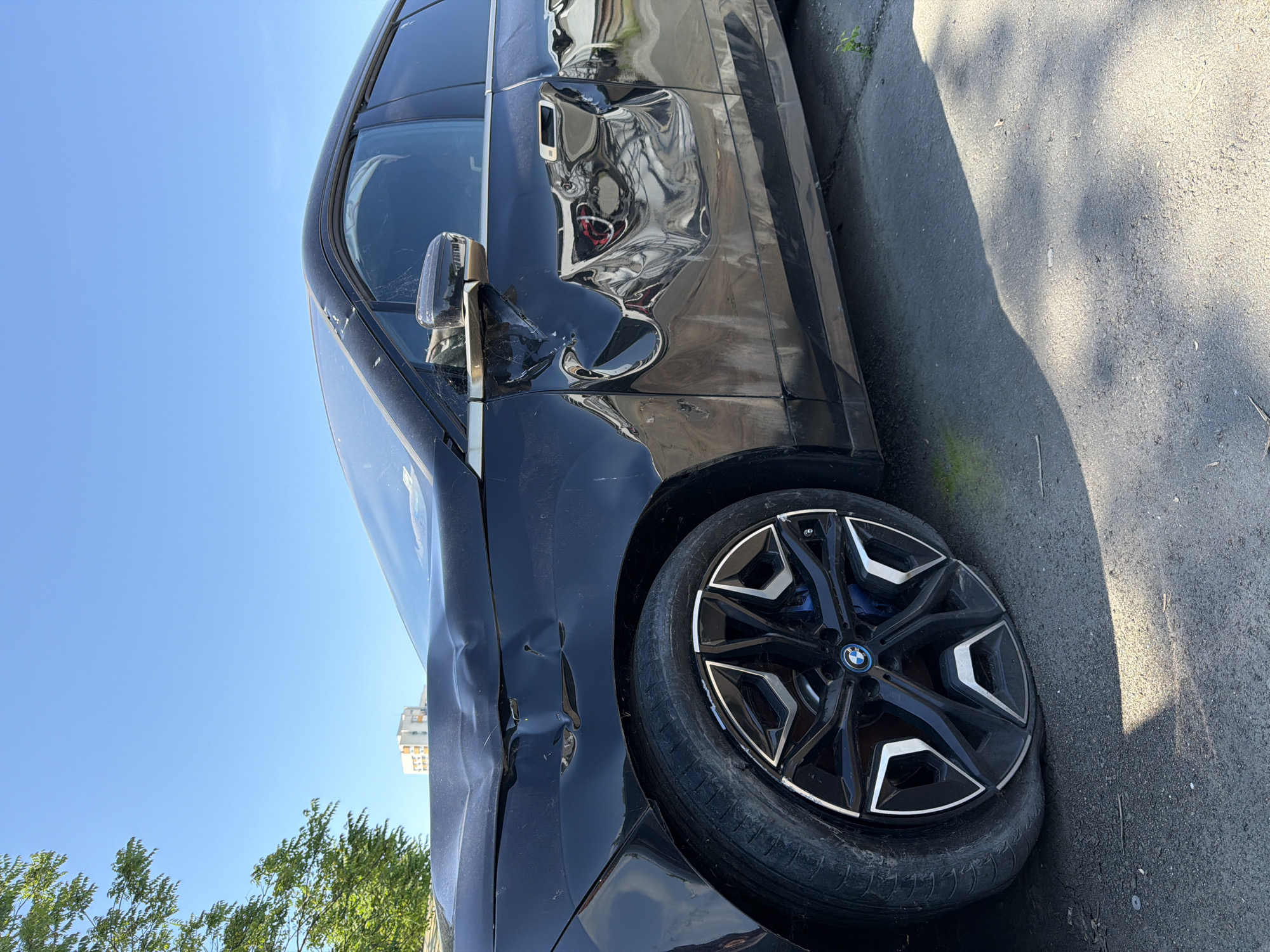 The image shows a damaged black BMW sedan. The front left side of the vehicle has significant damage, including dents in the fender and door, and a broken side mirror. The front left tire is visible and appears intact. The car is parked on a paved surface, likely a street or parking lot. The sky is blue and there are trees in the background, along with a building. The location is Burgas, Bulgaria.