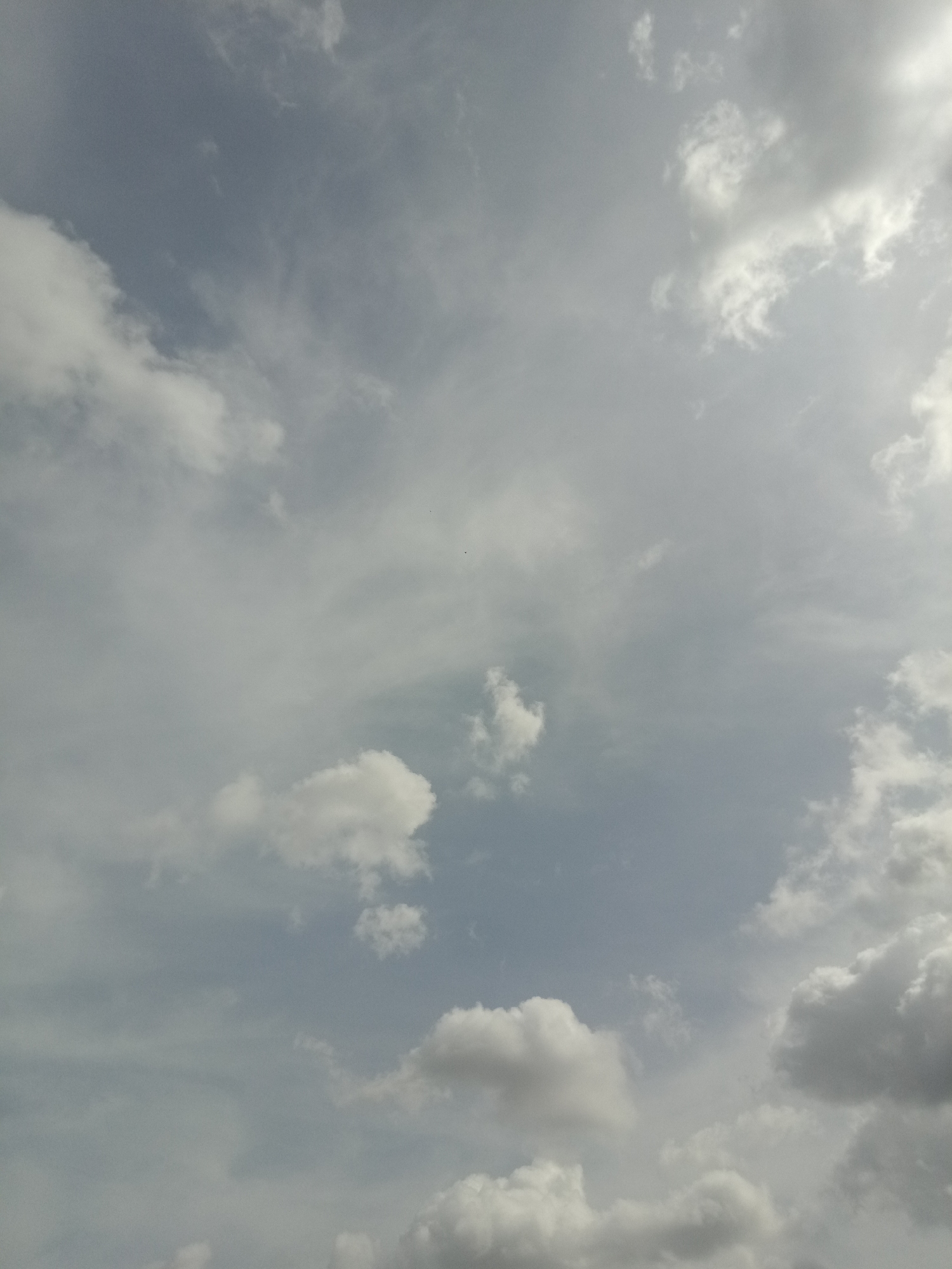 A wide-angle, high-angle shot captures a sky filled with clouds. The clouds vary in density and texture, with some appearing wispy and thin while others are more voluminous and opaque. The sky itself is a muted blue-grey, suggesting either an overcast day or the transition to or from sunset. The light source, likely the sun, is partially obscured by clouds in the upper right portion of the frame, creating a bright, hazy area. No buildings, people, or objects other than clouds are visible. The scene appears to be an outdoor environment, likely a city given the context of the prompt, with the sky dominating the frame.