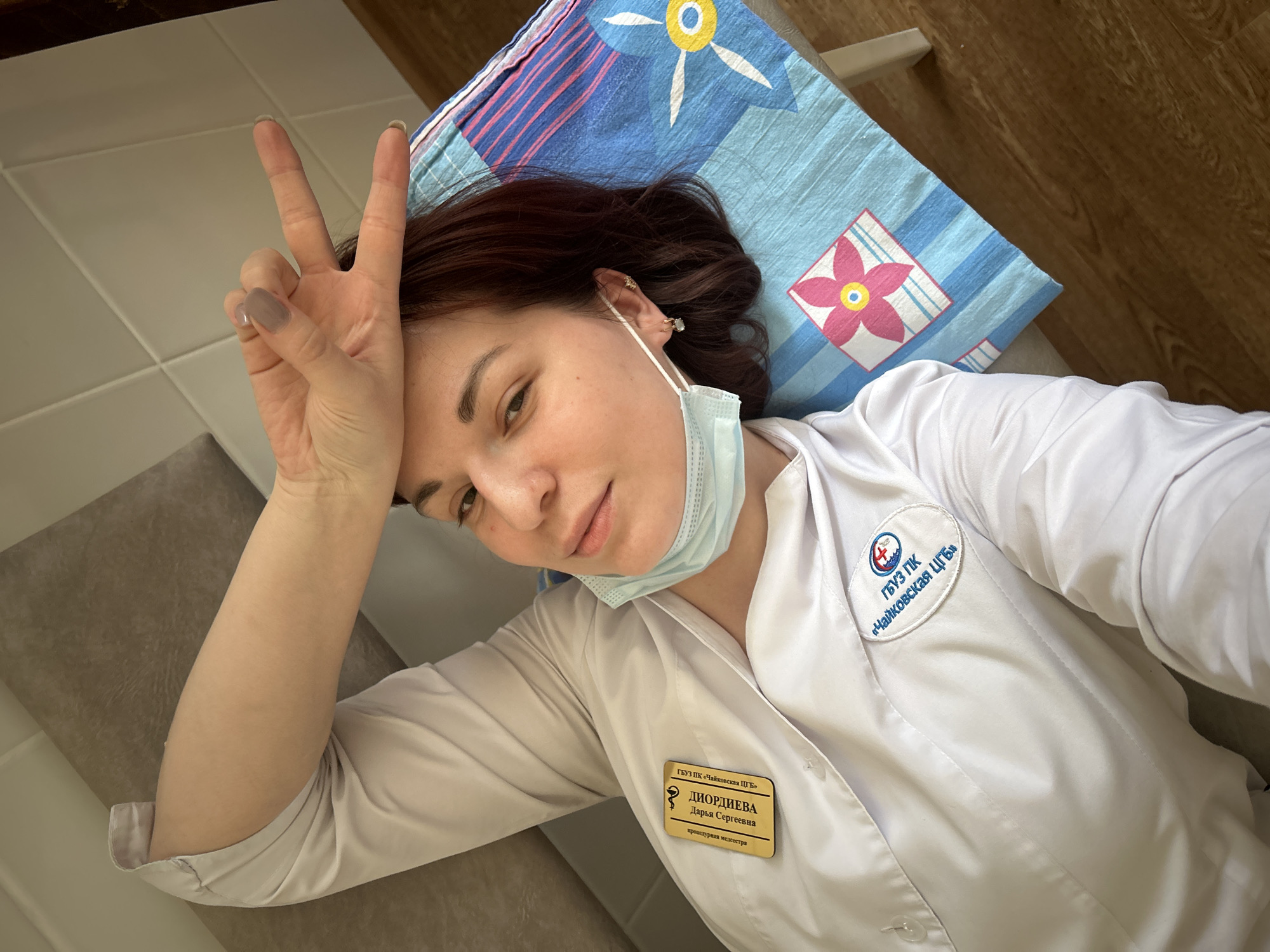 A young woman, appearing to be a medical professional, is captured in a selfie while lying down indoors, likely in Foky, Russia. She has brown hair and is wearing a white medical coat, with a light blue disposable face mask pulled down below her chin. Her left hand is raised in a peace sign near her head, and she has a relaxed expression with a subtle smile.
She is resting her head on a blue and white striped pillowcase adorned with yellow and pink floral patterns. The background includes light-colored surfaces and a section of what appears to be a wooden floor or wall on the upper right.
Pinned to her medical coat is an ID badge featuring Cyrillic text. It identifies her as "ДИОРДИЕВА Дарья Сергеевна" (DIORDIEVA Darya Sergeevna) and states her position as "процедурная медсестра" (procedural nurse). Both the badge and a circular patch on her left shoulder display the logo and name "ГБУЗ ПК «Чайковская ЦГБ»" (State Budgetary Healthcare Institution of the Perm Krai "Chaykovskaya Central District Hospital").