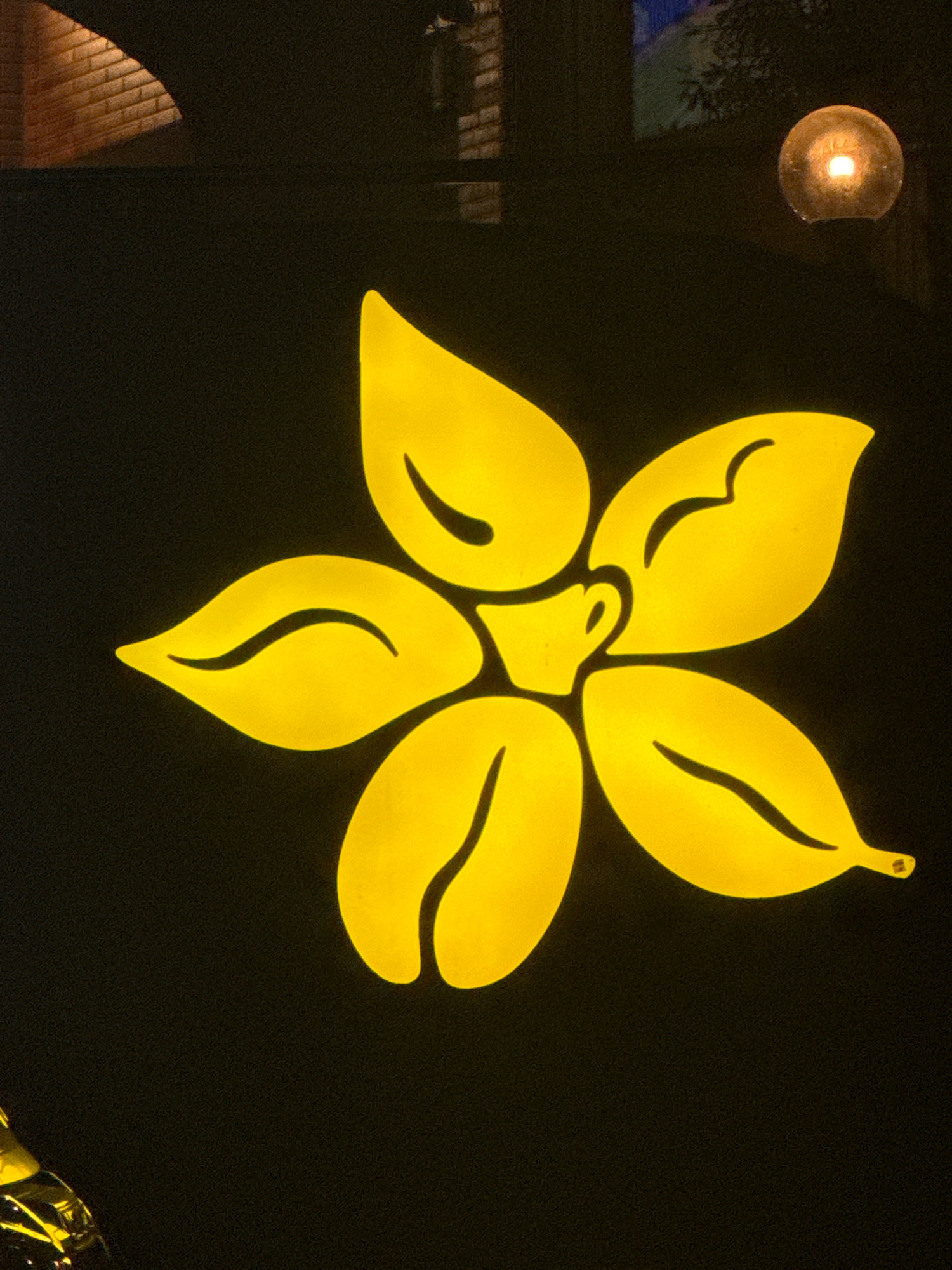 A backlit yellow flower logo is displayed on a dark, possibly black, background. The flower design features five prominent petals and a central element that resembles a coffee cup with a handle. The petals have subtle, darker yellow lines suggesting veins or contours. To the upper left of the flower, a section of a brick wall is visible, illuminated by a light source that casts a diagonal shadow. In the upper right quadrant of the image, a spherical light fixture with a warm glow is present. The bottom left corner shows a blurry, reflective object with yellow accents, possibly part of a display or signage. The scene appears to be an exterior shot, possibly a storefront or cafe in Agios Tychona Community, Cyprus, given the lighting and signage.
