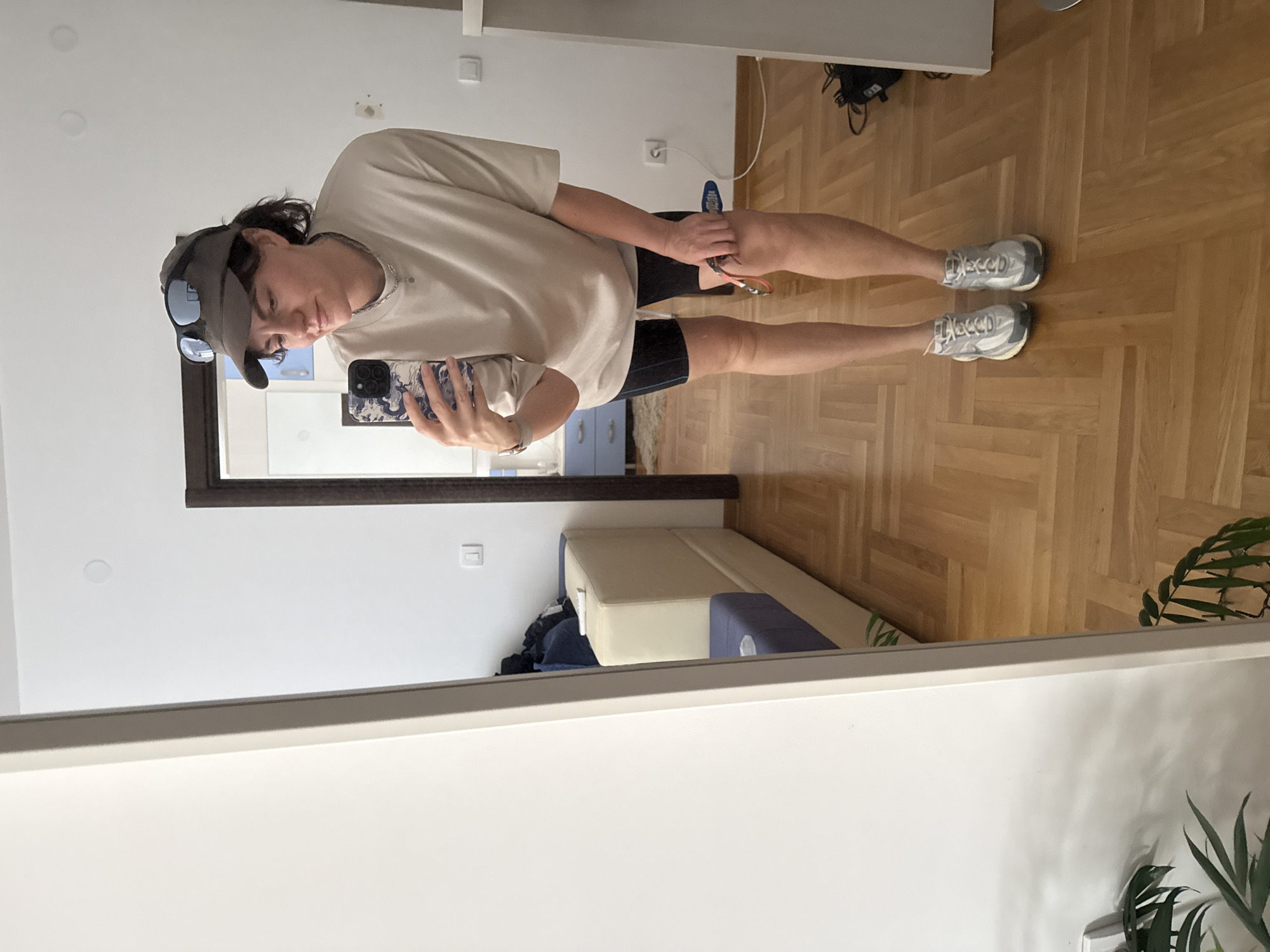 A person is standing in a room and taking a mirror selfie. The person is wearing a beige oversized t-shirt, black athletic shorts, silver running shoes, and a grey visor with sunglasses pushed up on top. They are holding a smartphone in their right hand and a blue and red object, possibly a hair brush, in their left hand. The room has light brown wooden parquet flooring and a white wall. To the left, a large mirror reflects the scene. In the background, a portion of a beige and blue sofa is visible, along with a closed wooden door. A plant with dark green leaves is also present in the lower left corner of the image. The image was taken in the city of Boreti, Montenegro.
