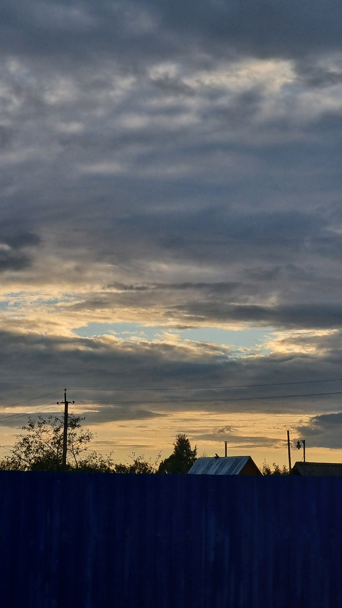 The image shows a cloudy sky at dusk over the city of Vysokoe, Russia. In the foreground, a dark blue fence runs horizontally across the bottom of the frame. Behind the fence, the silhouettes of trees and houses are visible against the muted tones of the sunset sky. The sky is filled with numerous horizontal cloud formations, with a break in the clouds revealing a lighter blue sky with hints of yellow and orange light. Power lines and utility poles are also visible in the mid-ground, breaking up the silhouette of the trees. The overall impression is one of a quiet, rural evening.