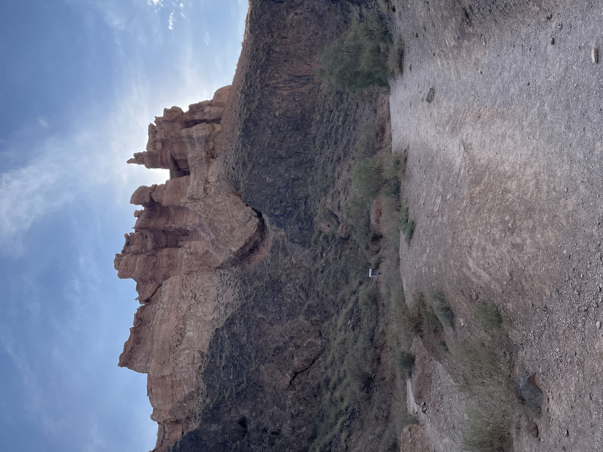 A wide, dusty path leads into a canyon. Towering rock formations, streaked with shades of red and dark brown, rise on either side. The sun shines through a hazy sky, casting a bright glow on the upper rock faces. Sparse, dry vegetation dots the canyon floor and clings to the rocky slopes. A small, white signpost stands near the base of the darker rock formation. The overall scene depicts a remote, arid landscape in Tamerlik, Kazakhstan.