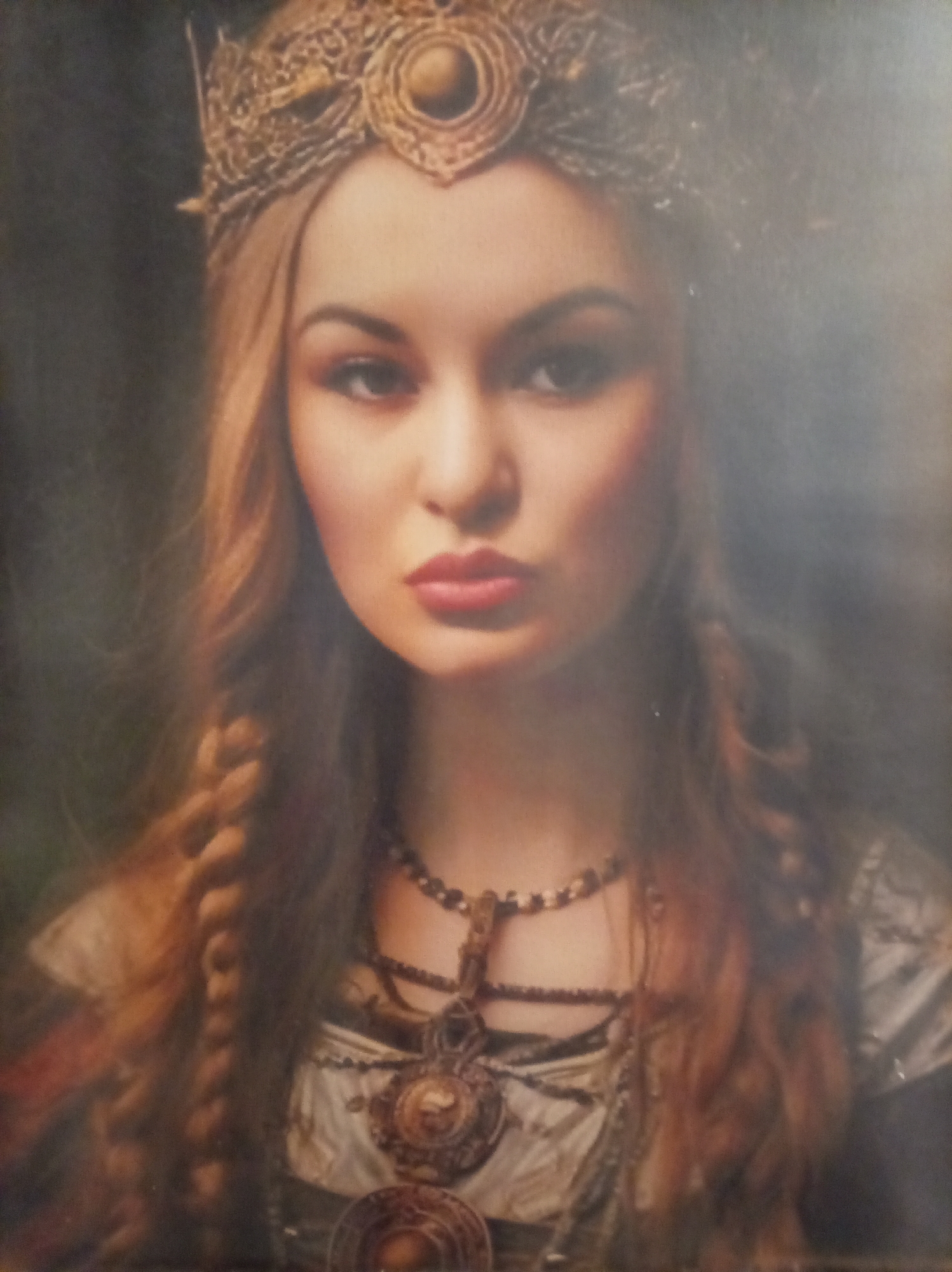 The image shows a close-up portrait of a young woman wearing an ornate golden crown and elaborate necklaces. Her hair is long and braided, with strands falling around her face and over her shoulders. She has fair skin, prominent cheekbones, and full lips painted with red lipstick. Her eyes are dark and her gaze is directed towards the viewer. The background is dark and out of focus, suggesting a studio or staged setting. There is no visible indication of location within the city of Yessentuki, Russia, or any context related to that city.