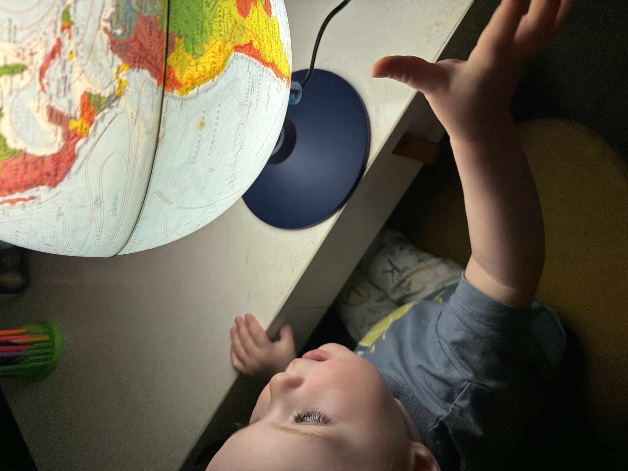 A child lies on their back, gazing up at an illuminated globe on a desk. The child is wearing a grey t-shirt and lies next to a pillow with a "K" pattern. The child reaches toward the globe with their right hand. A container of colored markers is visible to the left of the desk. The city of Votkinsk, Russia, cannot be determined from this image.