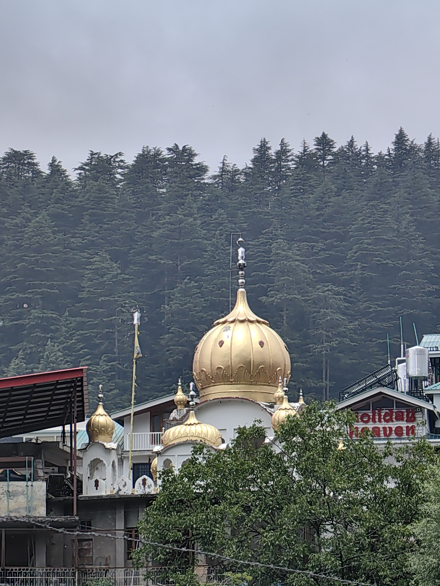 In Manali, India, a golden-domed building, possibly a temple or gurdwara, stands prominently against a backdrop of dense pine trees. The building features intricate architectural details, including smaller golden domes and white walls. To the right of the main structure, a building with a red sign that reads "Holiday Heaven" is visible, suggesting it is a hotel or resort. The scene is partially obscured by lush green foliage in the foreground. The sky is overcast.