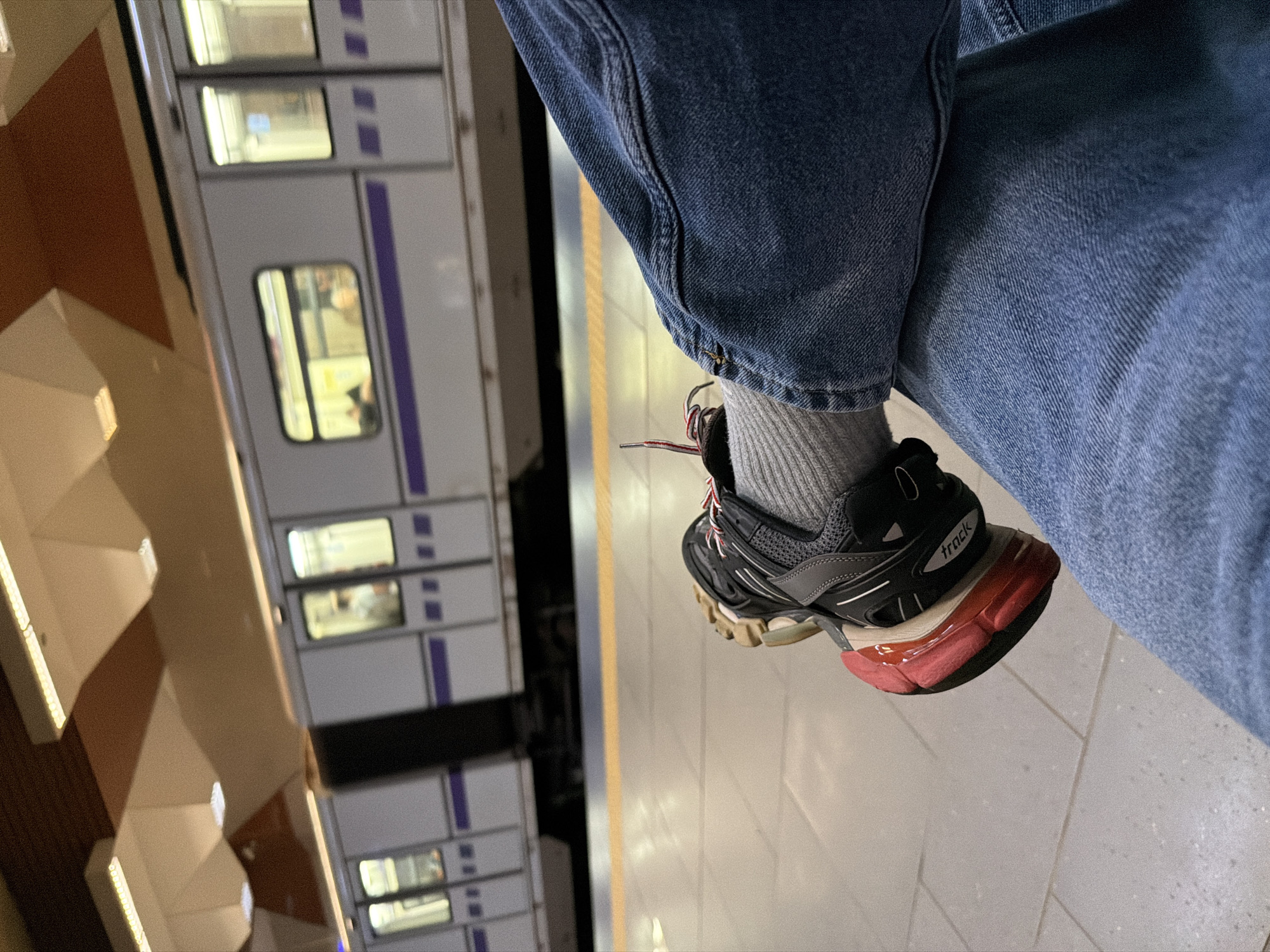 The image shows a person sitting with one leg crossed over the other on a platform, presumably in the Sofia Metro in Sofia, Bulgaria. The person is wearing blue jeans, gray socks, and black and gray "Track" brand sneakers. A metro train with closed doors is visible behind the person. The platform is tiled and has a yellow safety line. A brown wall with built-in lighting is visible on the left.