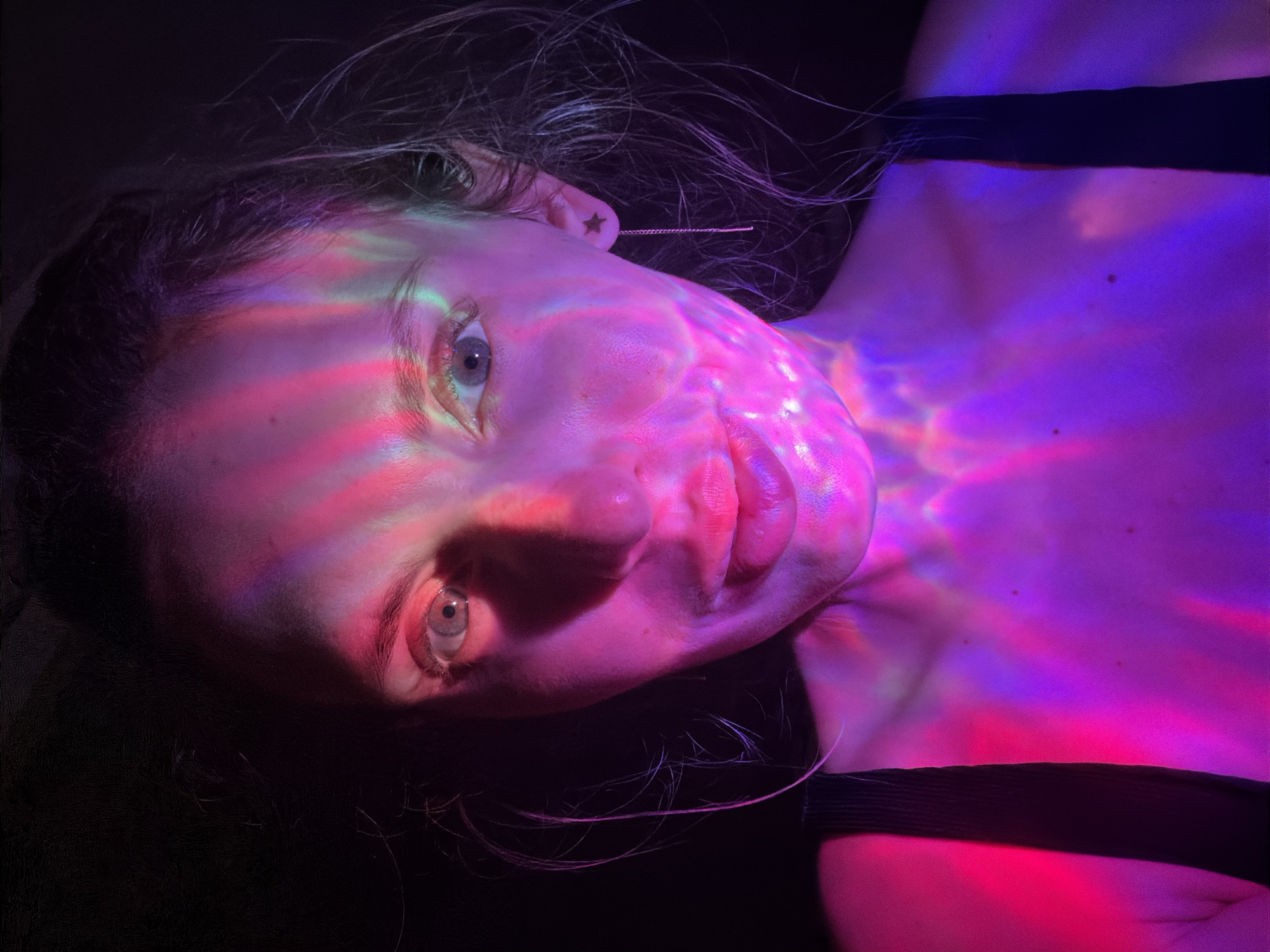 A close-up, dramatically lit portrait of a fair-skinned person, possibly female, likely taken indoors. The face is angled, with the person looking directly at the camera. The face is illuminated with pink, purple, green, and red lights, creating a pattern across the skin. The hair appears light brown or blonde, and is partially visible against the dark background. The person is wearing a black top with straps visible. The left ear is adorned with a small star earring with a hanging chain. The background is predominantly dark. There is no information to indicate that the photo was taken in Budva, Montenegro.