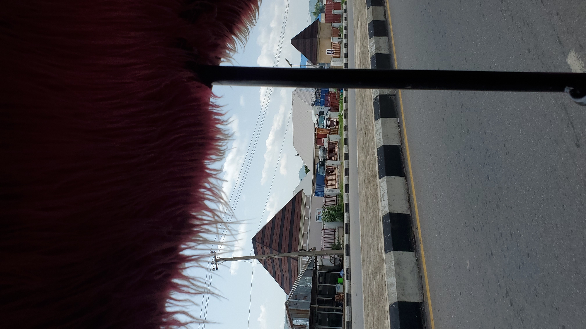 The image captures a street scene in Jalingo, Nigeria, viewed from inside a vehicle, with a prominent fuzzy red object framing the top of the frame. The foreground consists of a gray asphalt road with a yellow line and a curb marked with alternating black and white rectangular blocks. Beyond the curb, a paved area with similar black and white markings leads to a sidewalk and then a row of buildings.
The buildings vary in design. On the left, there's a structure with a corrugated metal roof and open-fronted sections, appearing to be a shop or market stall. Next to it is a house with a distinctive pyramidal roof covered in brown and black patterned shingles. Further down the street, other buildings are visible, including a larger structure with a light-colored roof and what appears to be shipping containers or storage units behind a metal fence. Another building to the right also features a pyramidal roof. Electrical poles and overhead wires are visible. Several vehicles, including what look like trucks, are parked or moving along the street. The sky is partly cloudy and blue, with hints of distant hills or mountains in the background. A single person is visible sitting near the open-fronted structure on the left.