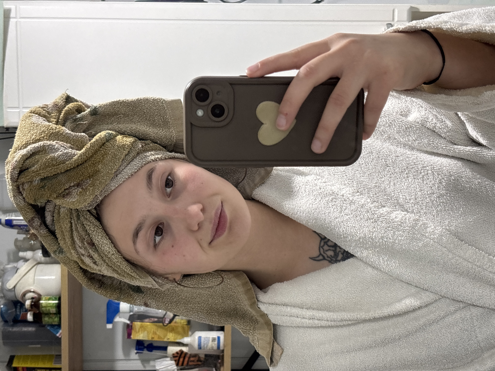 A selfie was taken by a young woman with a fair complexion. She is wearing a white bathrobe and has a towel wrapped around her head. Her phone, with a brown case and a heart-shaped design on the back, is held up to take the picture. The background reveals a cluttered bathroom with various items on shelves and a white cabinet. There is a tattoo visible on her collarbone. The location is not Фоки, Russia.