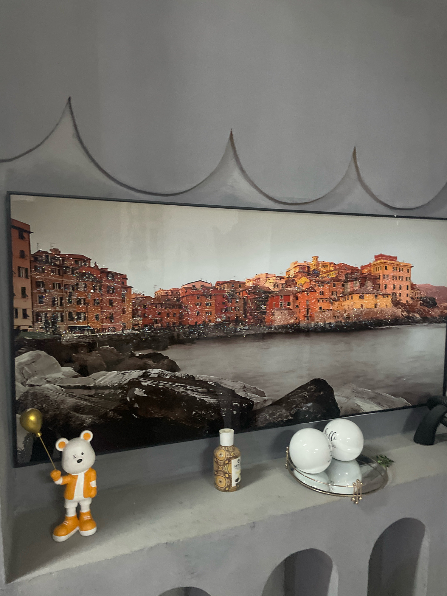 The image shows a wall with a large framed artwork depicting a coastal city with colorful buildings and a body of water. The artwork is positioned above a shelf. On the shelf, there is a small figurine of a bear holding a gold balloon, a bottle with an ornamental label, a small oval mirror, and two white spherical objects. One of the spherical objects has the word "hello" written on it. The wall above the artwork has a decorative scalloped pattern. The scene appears to be an indoor setting, possibly a home or office. There is no indication of the city of Ibadan, Nigeria.