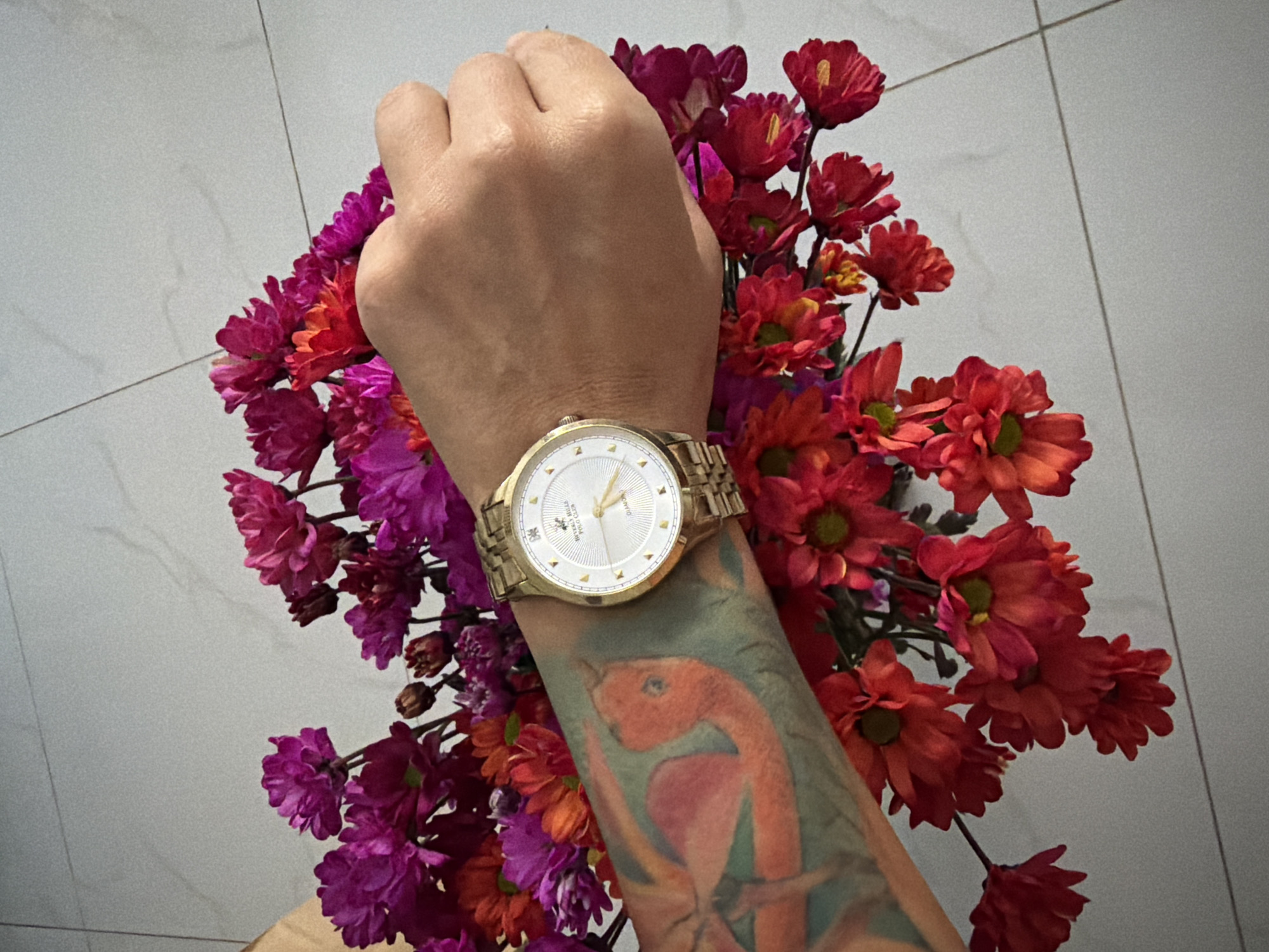 A close-up, top-down view shows a person's forearm with a gold wristwatch on the wrist, positioned against a bouquet of red and magenta flowers. The forearm has a large, colorful tattoo of a bird in orange and red hues. The flowers are arranged in a loose bunch, with some petals scattered. The background consists of a light-colored tiled floor. The scene is indoors, possibly in a residential setting in Nha Trang Ward, Vietnam.