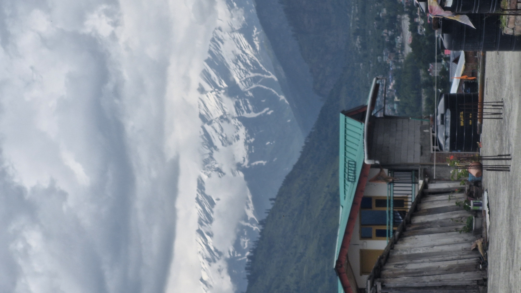 This image depicts a mountainous landscape with a small dwelling in the foreground. The dwelling has a green roof and appears to be made of wood and brick. In front of the dwelling, there are two black water tanks, one with text that reads "DIPLA IT SL". Several metal rods are visible sticking out of the ground in front of the water tanks.
The middle ground of the image shows a lush green mountainside dotted with trees. In the background, towering snow-capped mountains rise against a cloudy sky. The scene is likely a rural area in Vashisht, India, given the architecture and the natural environment. The lighting suggests it is daytime, possibly overcast. There are no people visible in the image.