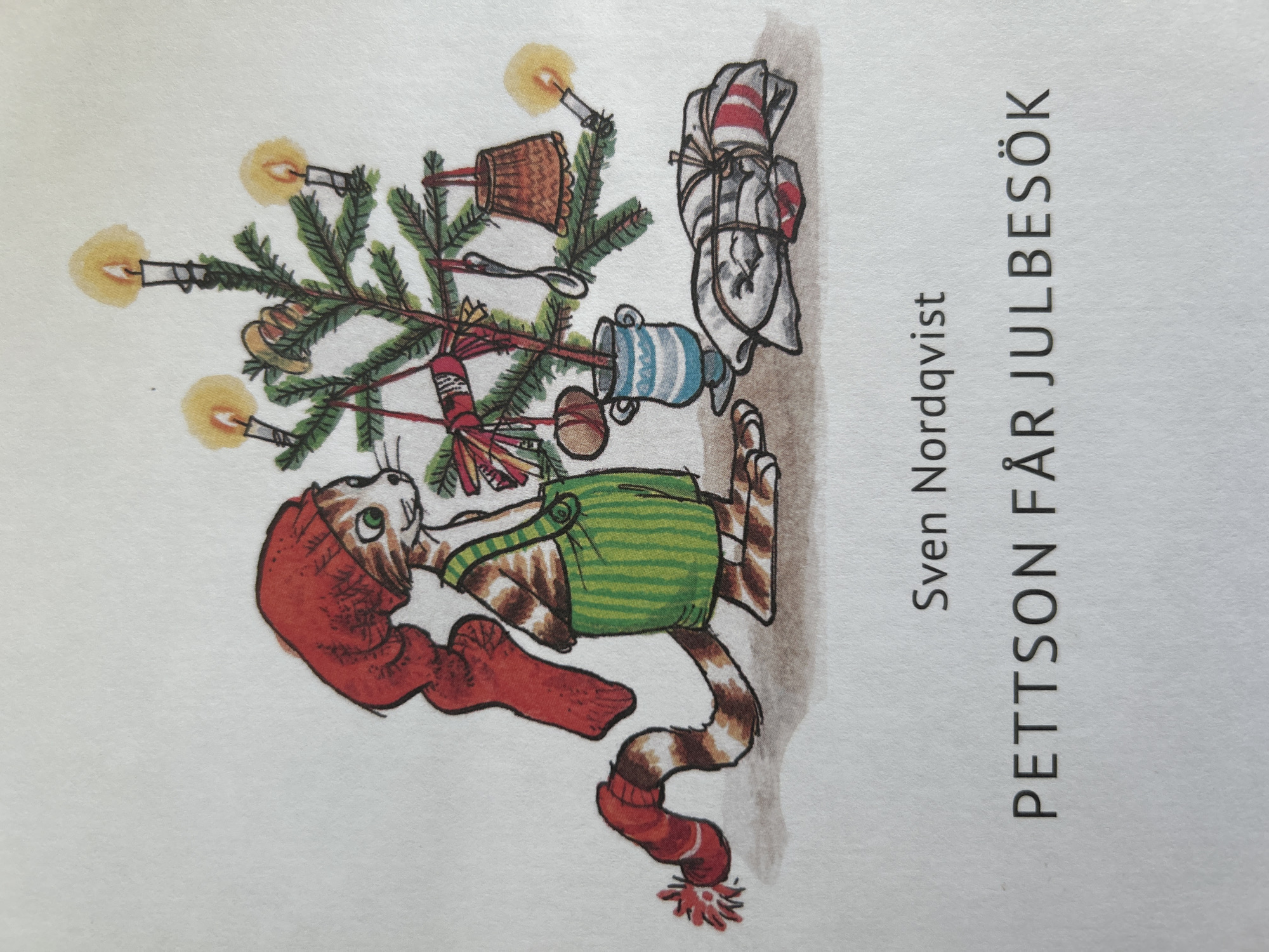 An illustration depicts a cartoon cat wearing a red hat and green overalls, standing next to a decorated Christmas tree. The cat has a striped tail adorned with a red knitted object. The Christmas tree is lit with candles and decorated with ornaments, including a small brown basket, a spoon, and what appear to be bundles of red and white fabric. A blue and white striped vase holds the tree. In the foreground, a bundle of wrapped gifts lies on the ground. Below the illustration, the text "Sven Nordqvist" is printed, followed by "PETTSON FÅR JULBESÖK". The scene is set against a white background.