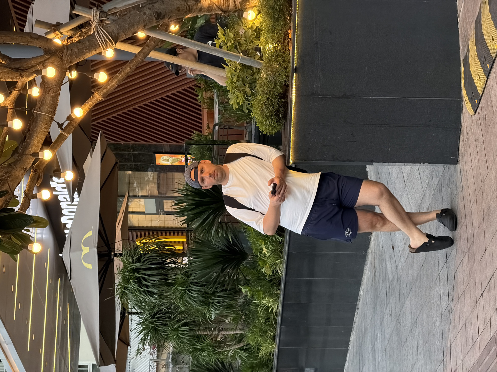 The image shows a person standing outside a McDonald's restaurant in Nha Trang Ward, Vietnam. The person, a man, is wearing a white t-shirt, navy blue shorts, a baseball cap, and black sandals. He is leaning against a dark-colored wall, holding a cell phone in his hand. Behind him is dense green vegetation. A McDonald's restaurant, with its logo prominently displayed, is visible in the background. Decorative lights are strung on a tree, contributing to a warm ambiance.