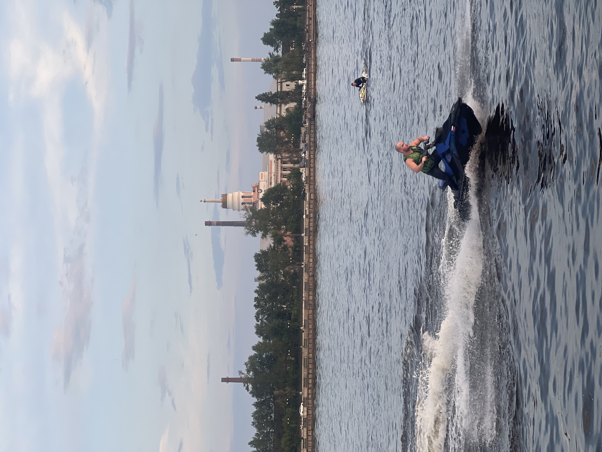 The image shows a man riding a jet ski on a body of water, with a factory in the background. The man is wearing a green life vest and appears to be moving at a fast speed, creating a large wake. In the distance, there are several industrial buildings with smokestacks, suggesting an industrial area of the city. Trees line the waterfront between the water and the buildings. The sky is partly cloudy, with a soft, diffused light. Another person on a jet ski is visible further in the background on the right. The overall scene depicts a recreational activity taking place near an industrial landscape. The image does not provide any specific visual cues to confirm the location as Izhevsk, Russia.