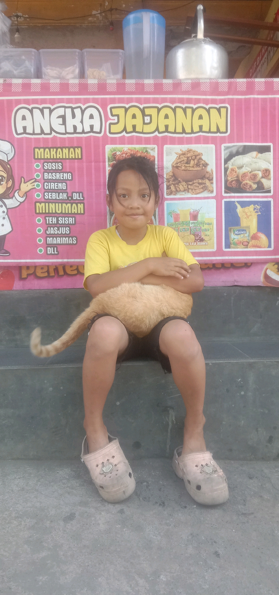 A young Indonesian girl, possibly of East Asian descent, sits on a concrete step in front of a pink banner advertising "ANEKA JAJANAN" (Various Snacks) in Palasari, Indonesia. The banner displays images of food and drinks, including fried foods and juices. The girl is wearing a yellow t-shirt and dark shorts, with light-colored slip-on shoes. A light-brown cat is perched on her lap, with its tail visible to the left. The background shows shelves with clear containers and a large blue water jug, suggesting a food stall or eatery. The overall scene appears to be outdoors, with natural lighting.