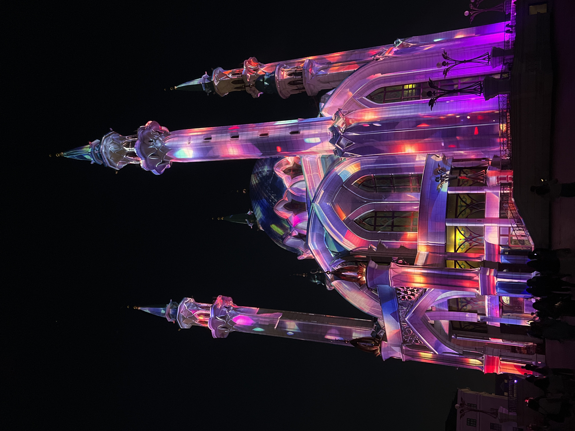 The image shows the illuminated Kul Sharif Mosque in Kazan, Russia, at night. The mosque features a central dome and four minarets, all adorned with multicolored lights. People are visible at the bottom of the image, and there are buildings in the lower right corner. The sky is dark.