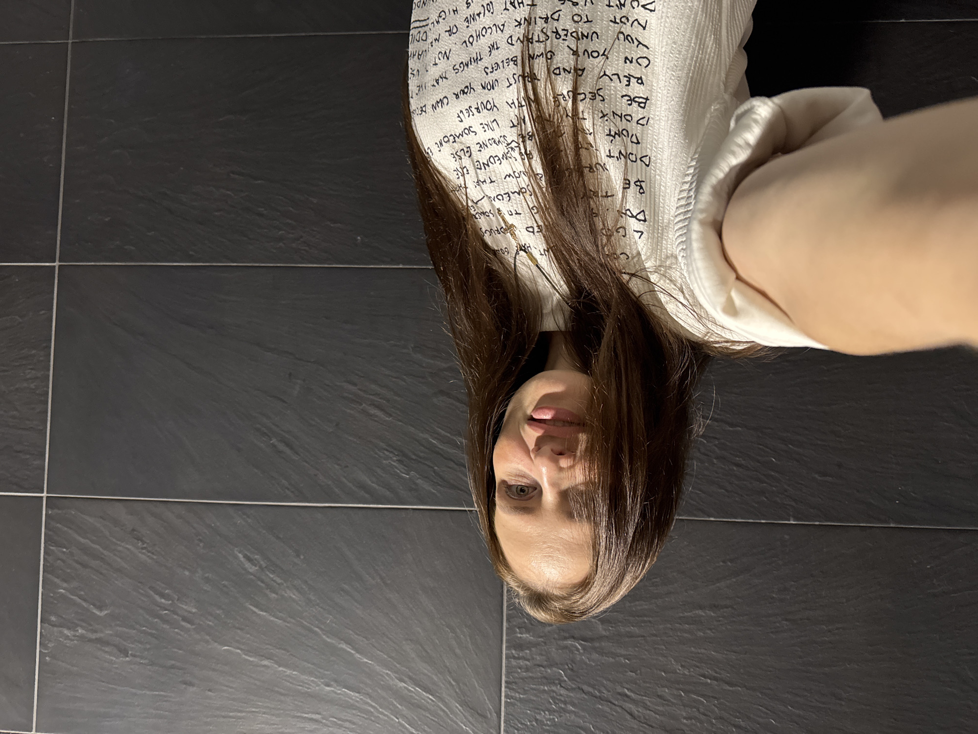 The image shows a woman lying on dark grey tiled flooring, looking at the camera. She has long brown hair that partially covers her face. The woman is wearing a white long-sleeved shirt with black text printed all over it. The text appears to be a series of statements, some of which are visible and legible, like "BE SECURE WITH YOURSELF", "DON'T TRY TO DRINK ALCOHOL", and "TRUST UPON YOUR OWN BELIEFS". The setting appears to be indoors, given the tiled floor and the lighting. The orientation of the image is sideways, with the woman's head at the bottom and her feet towards the top. The woman's expression is neutral, with her tongue slightly protruding. There are no clear indications of the time of day or weather. The location context provided is Chaikovsky, Russia.