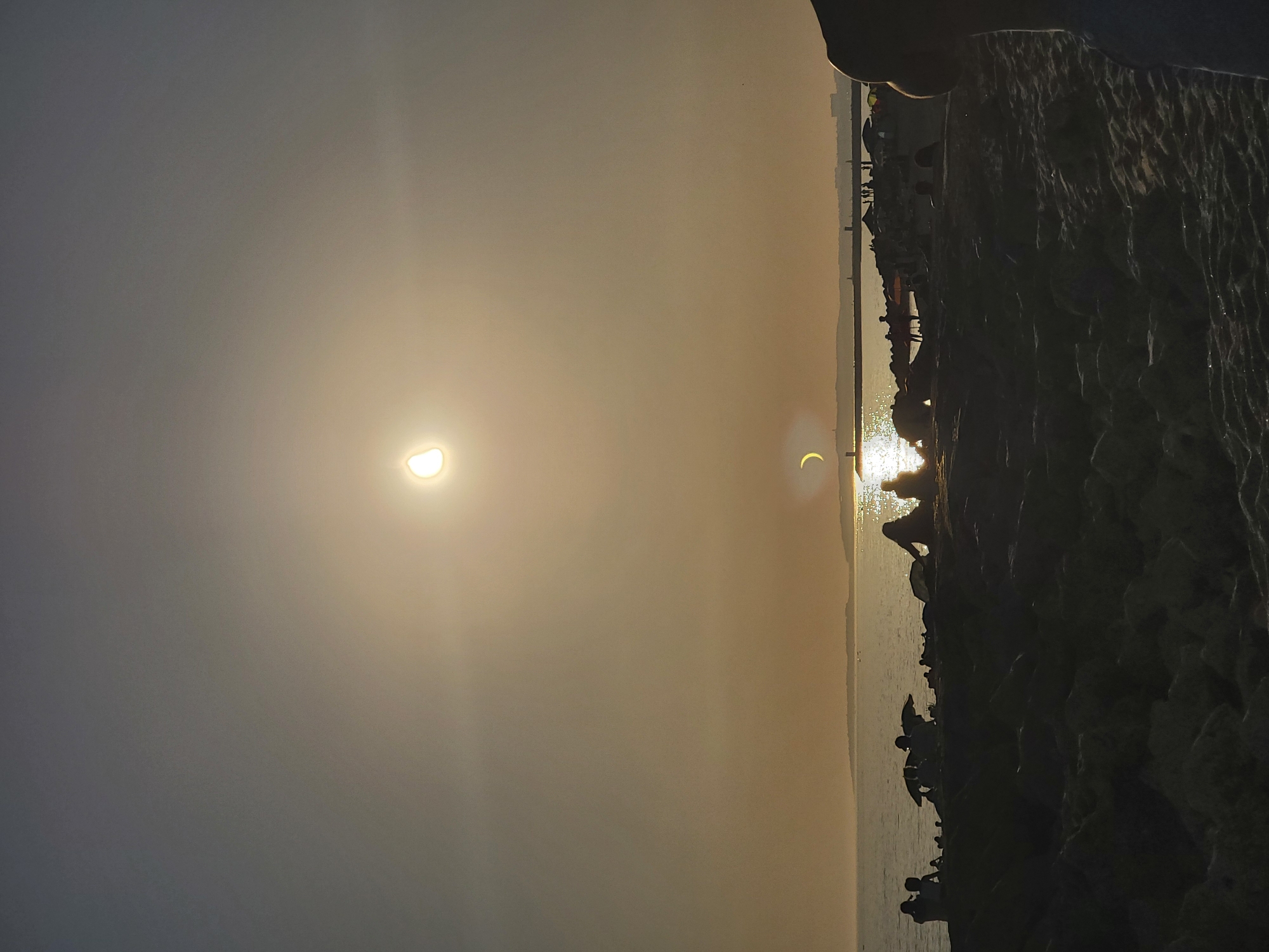 This image captures a scene at the beach in Quarteira, Portugal, during what appears to be a solar eclipse. The sun, partially obscured by the moon, is visible in the sky, casting a warm, hazy glow over the scene. Silhouettes of people are spread across the foreground, seated and standing on the sandy beach and a rocky breakwater. Some individuals are under beach umbrellas, suggesting a relaxed, recreational atmosphere. The calm sea stretches towards the horizon, where a distant landmass with buildings and a lighthouse can be seen. The overall mood is serene, with the natural phenomenon of the eclipse adding a unique element to the typical beach setting.