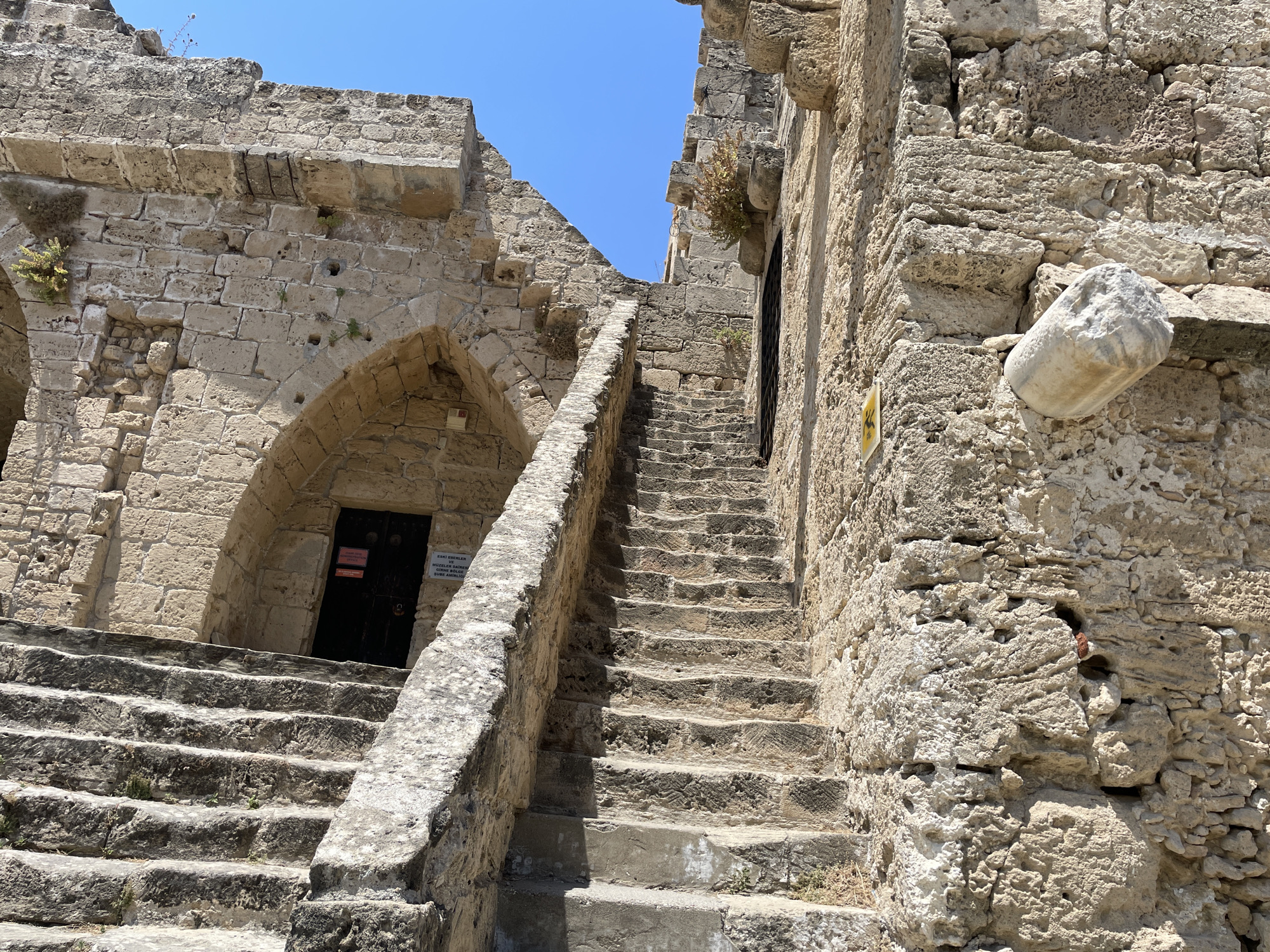 A stone staircase ascends between ancient stone walls under a clear blue sky. The stairs are weathered and uneven, with some modern concrete repairs visible at the bottom. The walls are constructed of rough-hewn stone blocks and show signs of age and erosion, with small plants growing in crevices. To the left, an arched doorway with a dark metal door is visible. To the right, the stone wall rises steeply, with a cylindrical stone fragment protruding from it. Signs are visible on the doorway and the wall, one of which appears to be in Turkish. This scene is located at Kyrenia Castle in Kyrenia, Northern Cyprus.