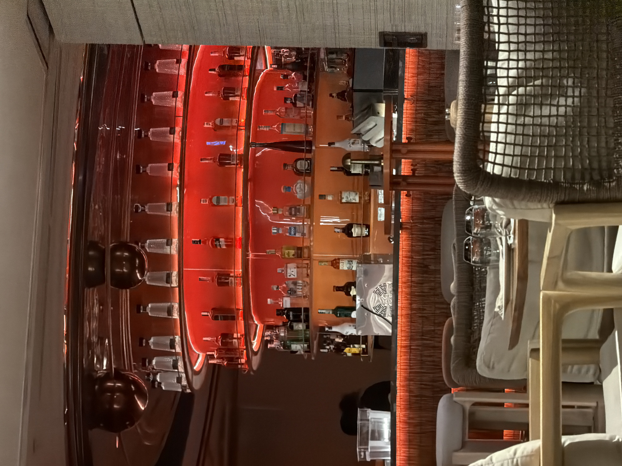 This is a dimly lit interior shot of a bar with a prominent display of liquor bottles. The bar area is characterized by warm, red ambient lighting emanating from behind the shelves of bottles and the counter. The shelves are stacked with numerous bottles of various shapes and sizes, suggesting a well-stocked bar. In the foreground, there are tables and chairs, some of which appear to be part of a dining or lounge area. Two people are partially visible in the background, one seated and the other in silhouette behind the bar, implying the bar is open for business. The architectural elements include textured wall paneling on the right side of the image and a reflective ceiling. The scene is consistent with a modern bar or restaurant establishment. This is the bar at the La Plage restaurant in Nha Trang, Vietnam.