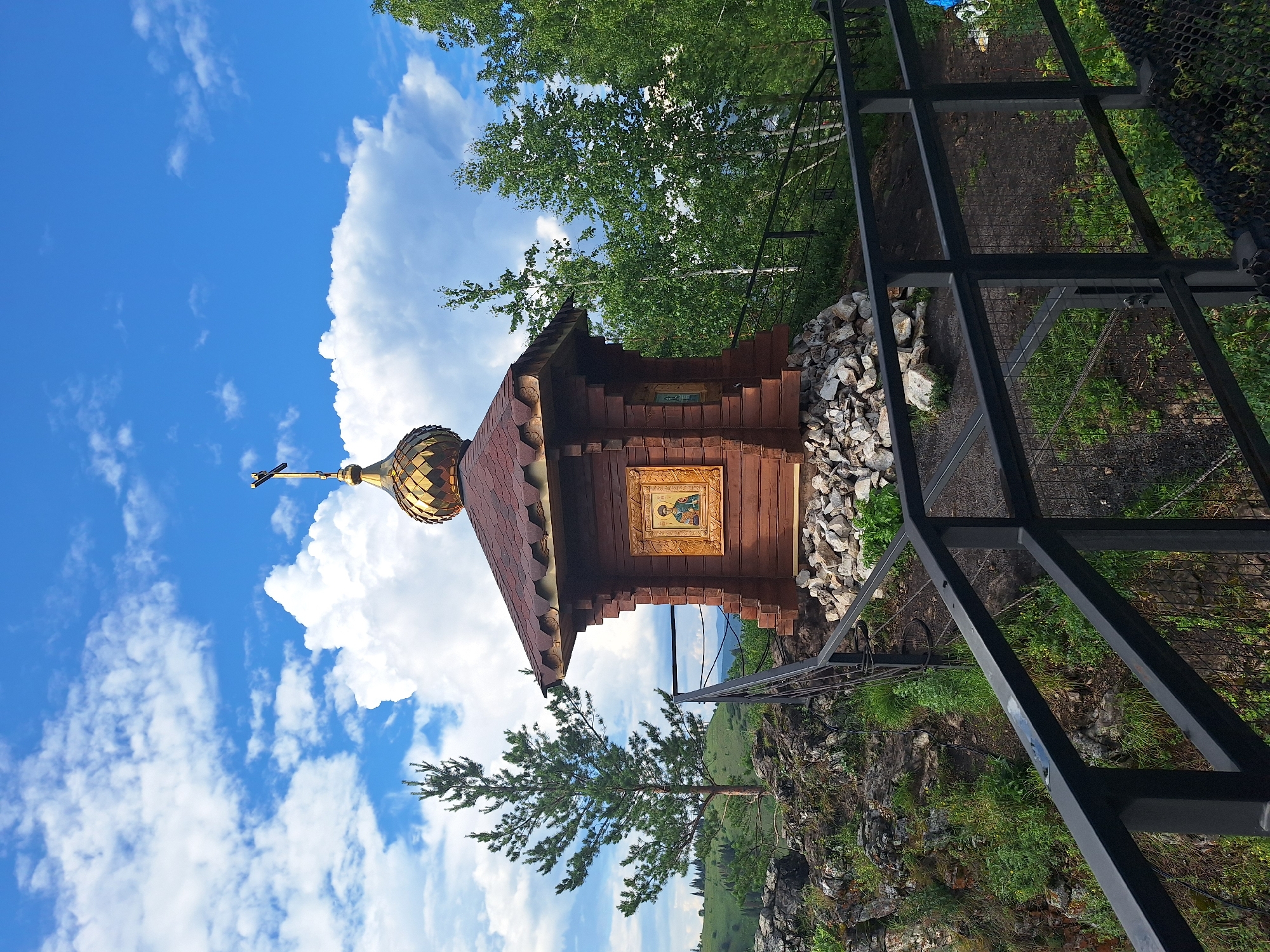 A small, wooden, Orthodox chapel with a golden dome stands on a rocky outcrop. The chapel features an icon in a gold frame on one side. A metal railing surrounds the area, leading towards the chapel. The background consists of lush green trees and rolling hills under a bright blue sky with scattered white clouds. The location is the "Skala Belaya" (White Rock) natural park in Sim, Russia.